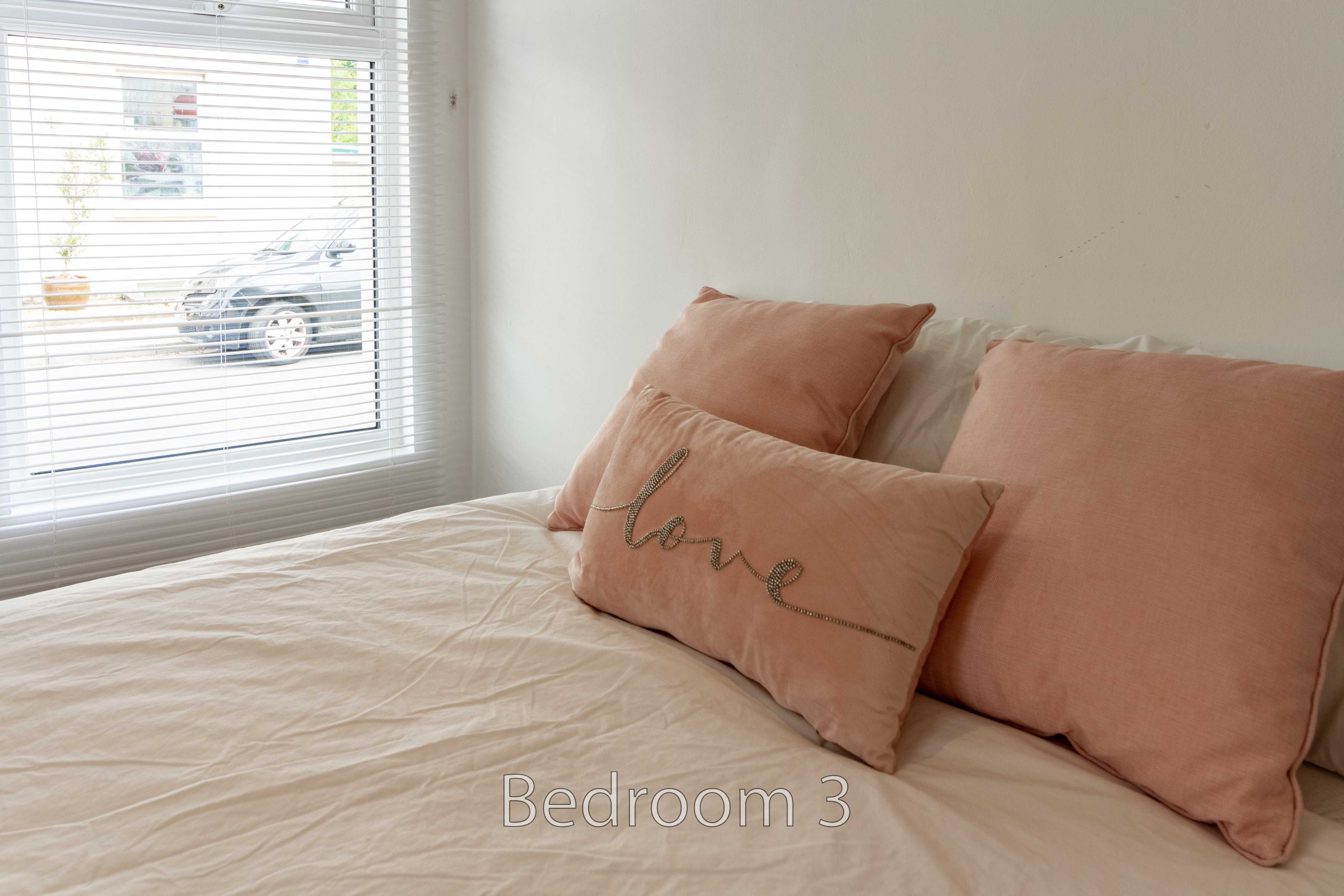 A bed with peach pillows and cushions beside a window with blinds.