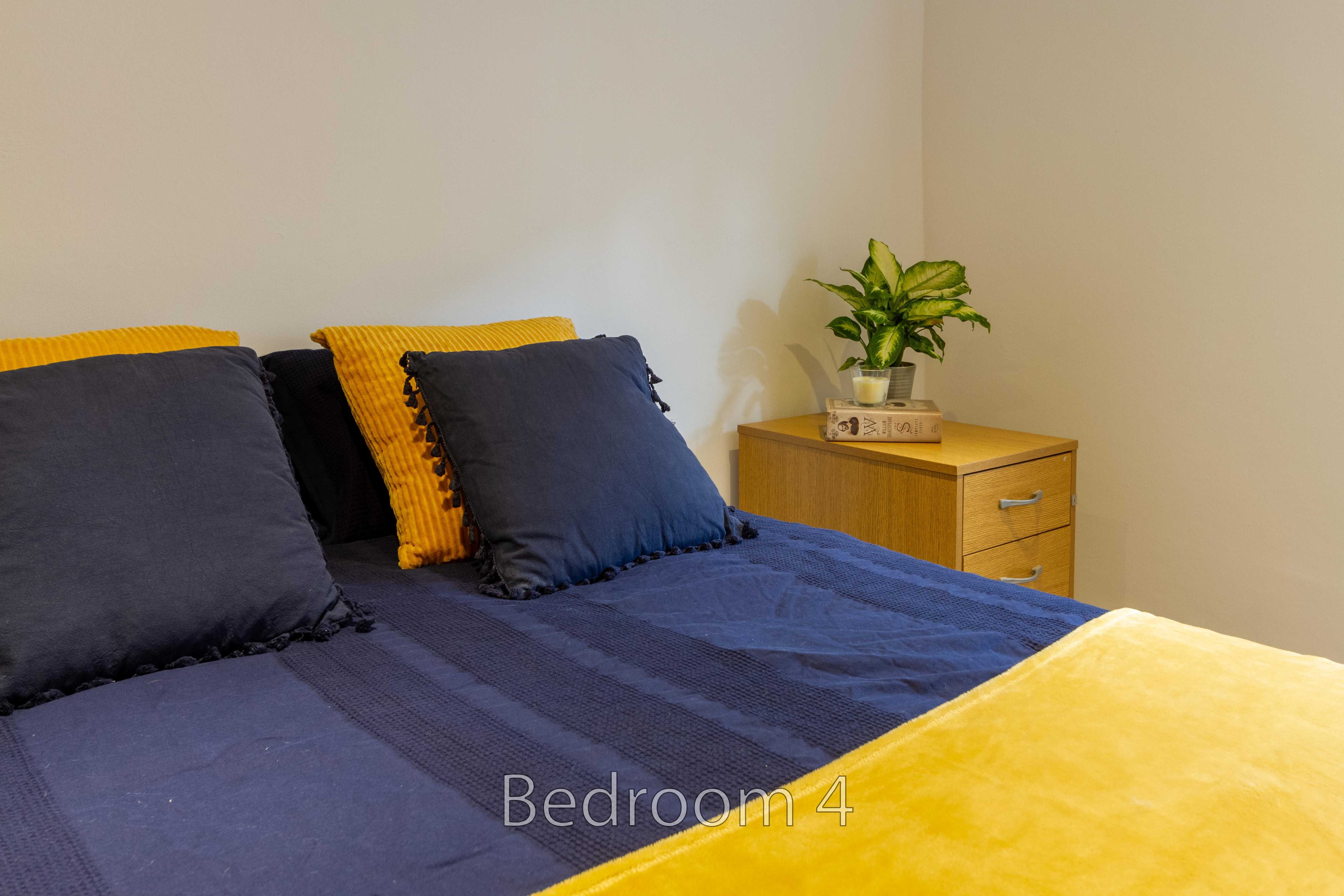 A bedroom with a neatly made bed, dark blue bedding, yellow cushions, and a wooden bedside table with a plant.