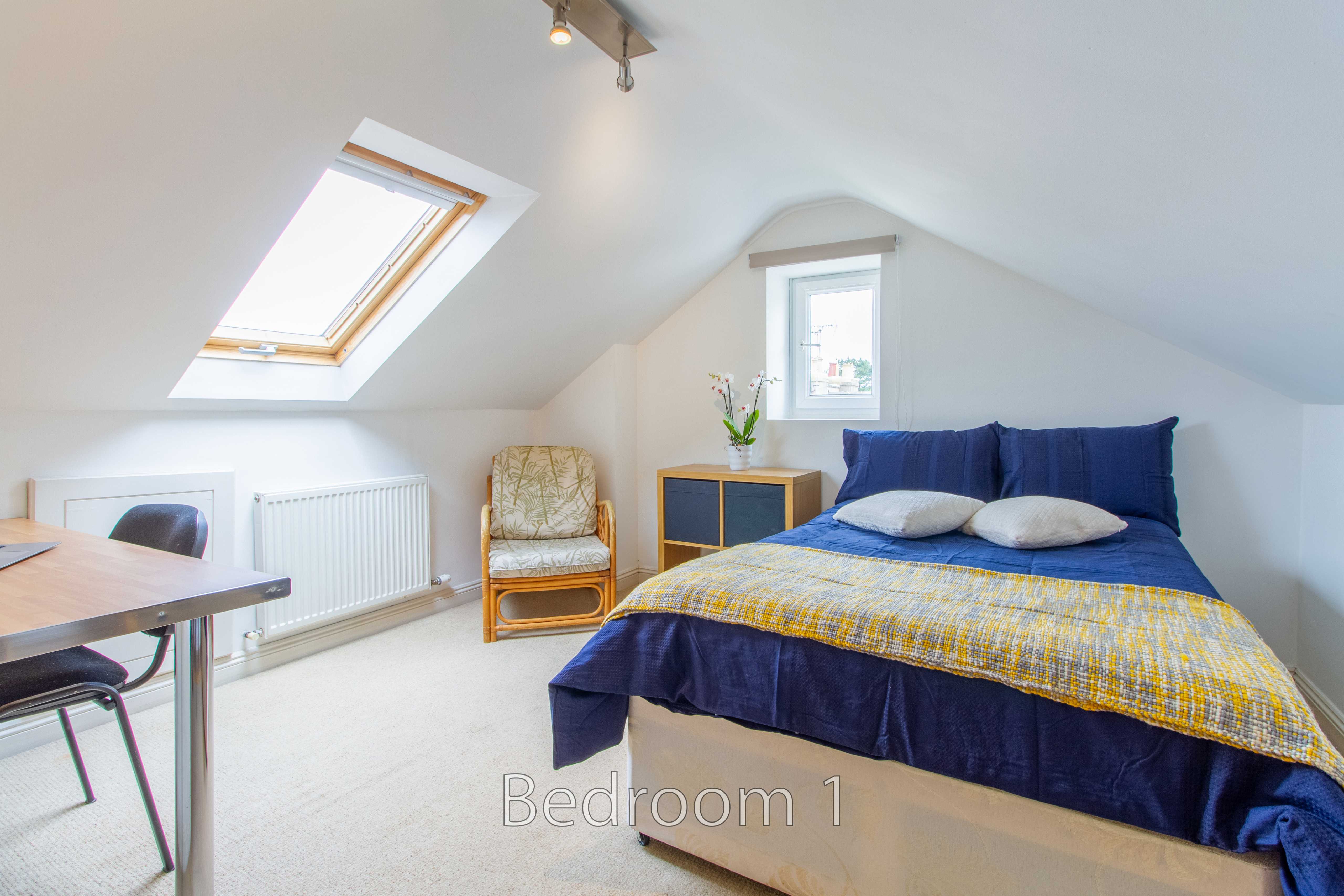 A furnished attic bedroom with a double bed, skylight, small window, chair, desk and radiator.
