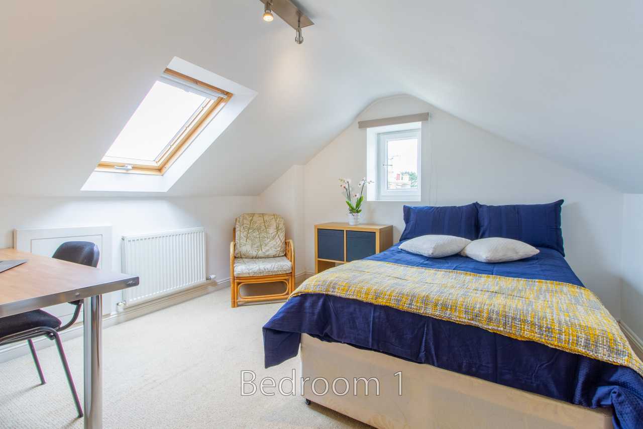 A furnished attic bedroom with a double bed, skylight, small window, chair, desk and radiator.