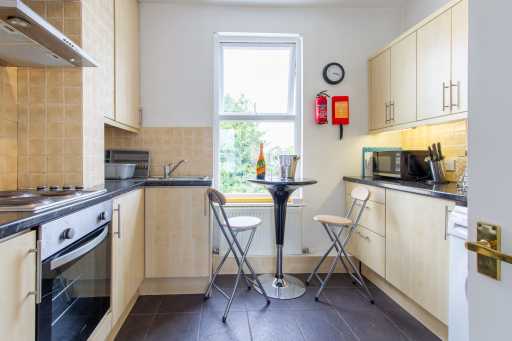 A bright shared kitchen with cabinets, cooker, sink, microwave, tiled walls and a small table with two folding chairs.
