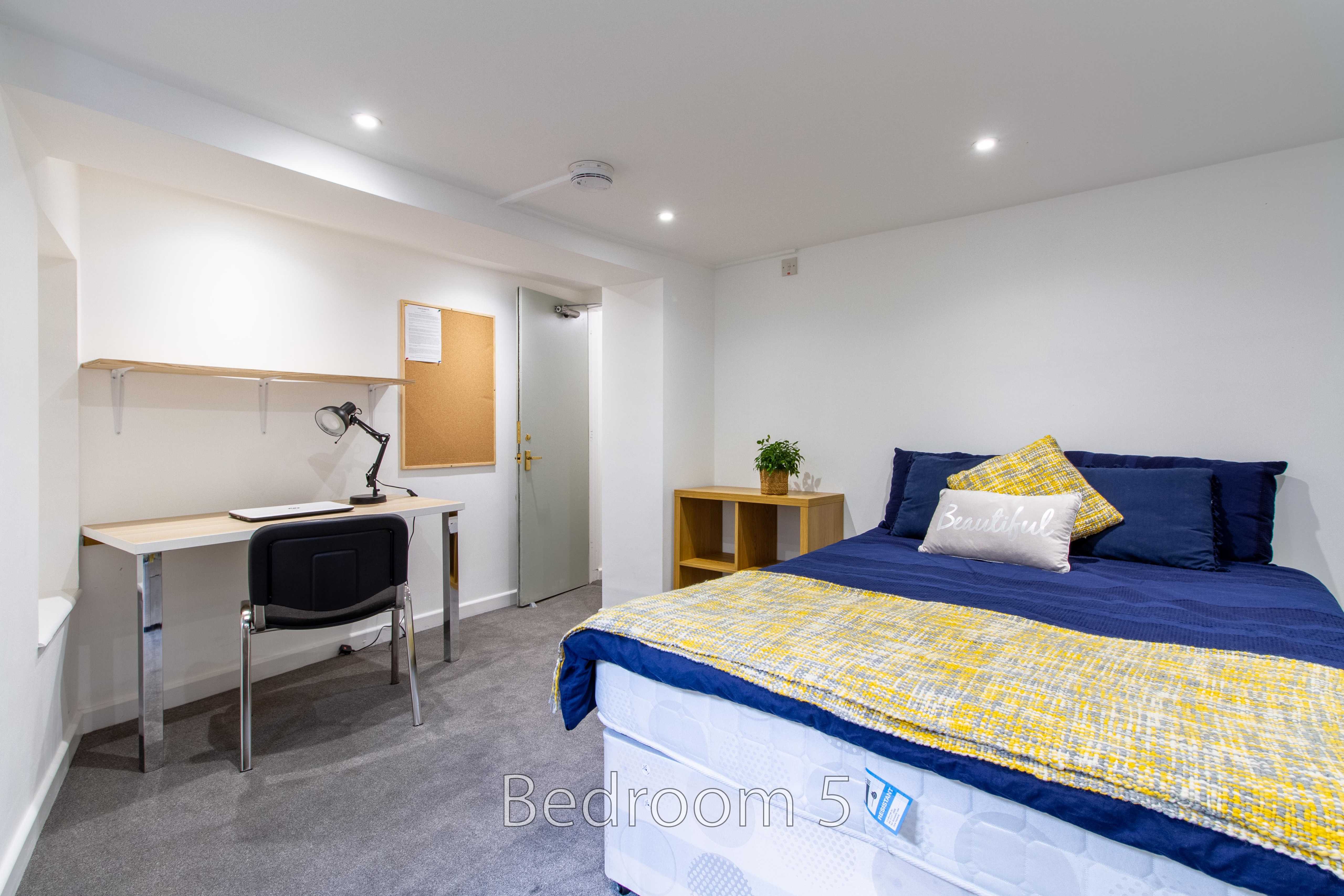 Furnished bedroom with a bed, desk, chair, shelving, and wall-mounted noticeboard.