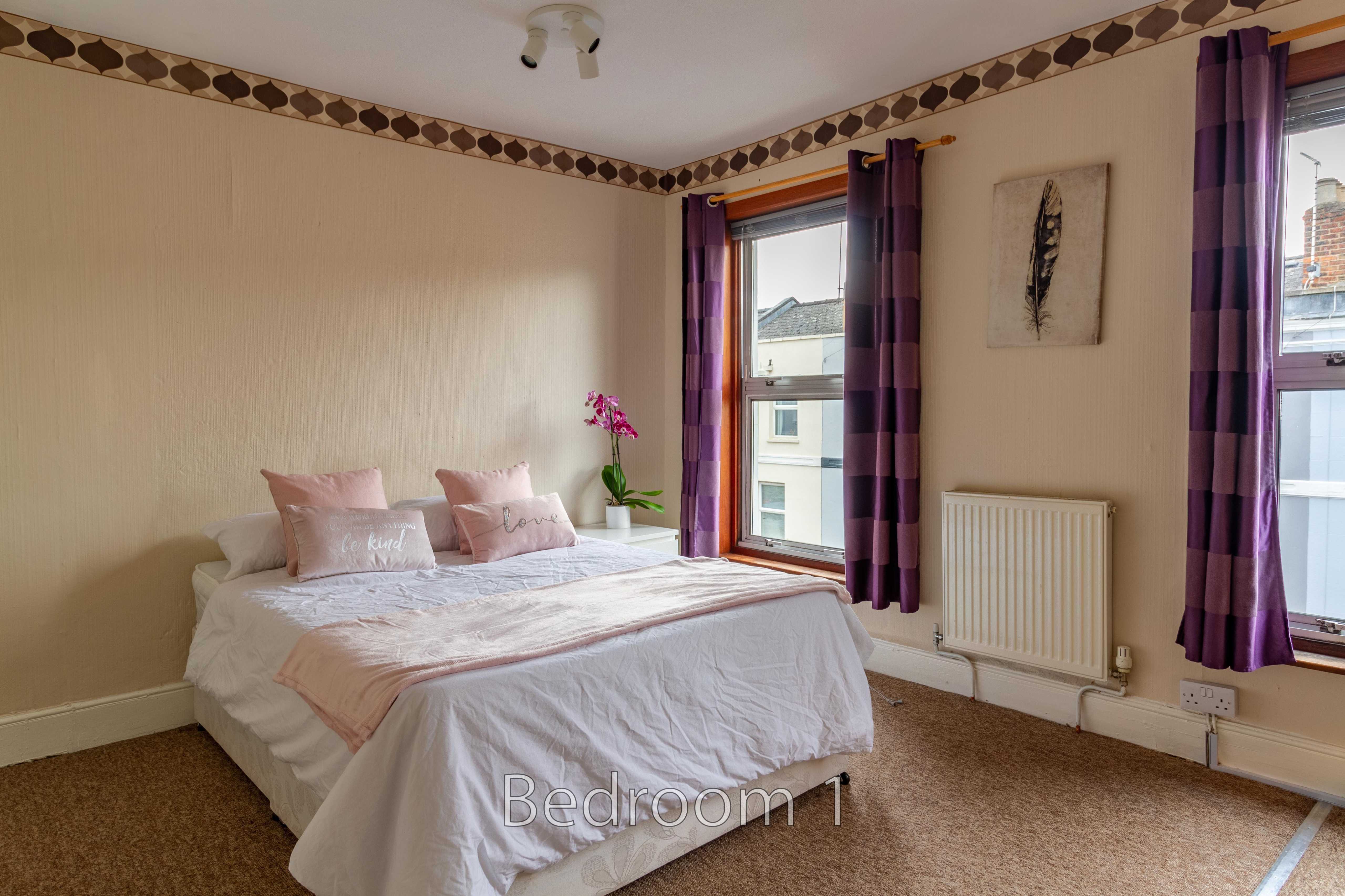 A furnished bedroom with a double bed, purple curtains, two windows, a radiator and wall decor.