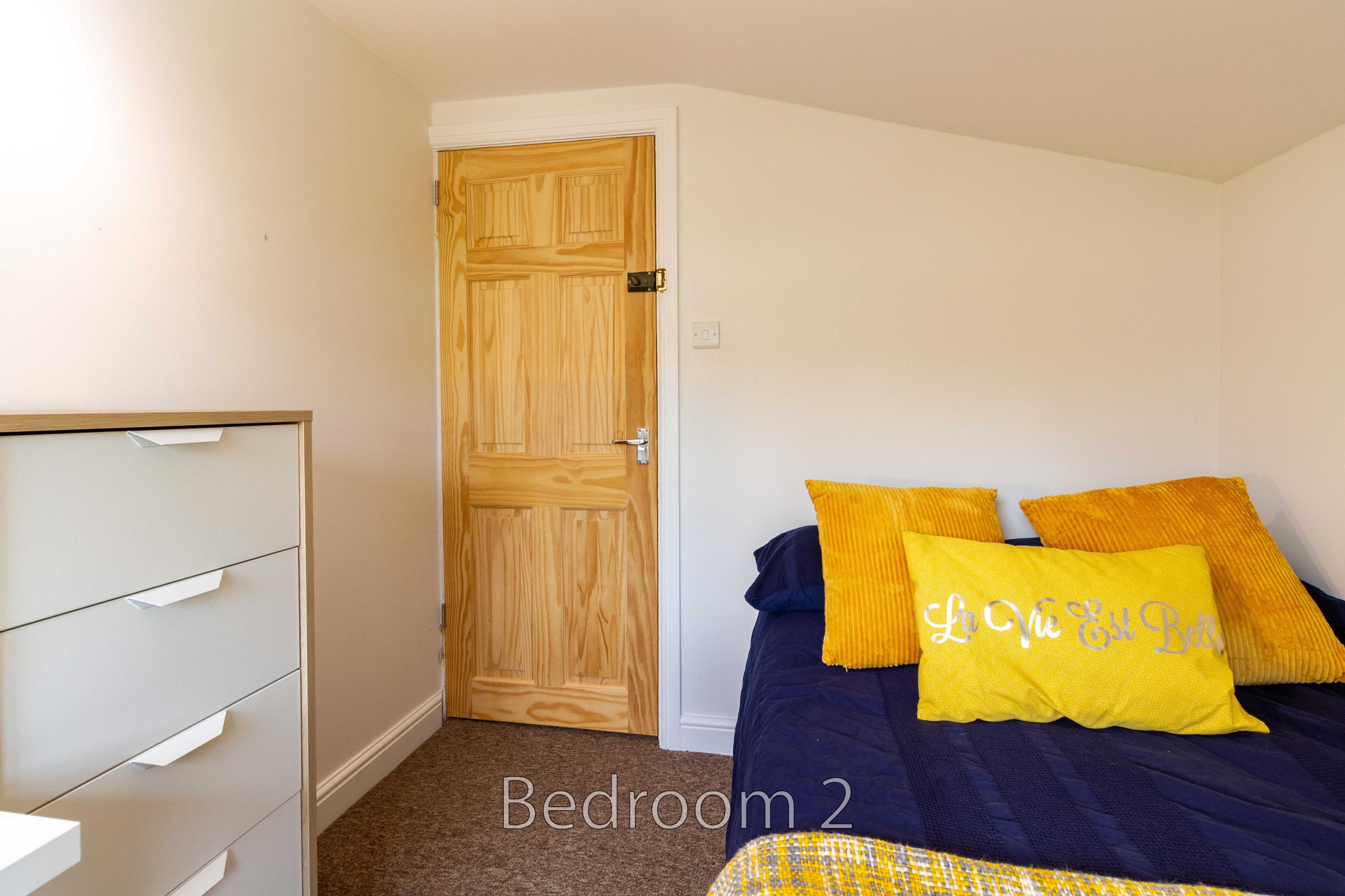 A bedroom with a bed, yellow and blue pillows, a wooden door, and a white chest of drawers.