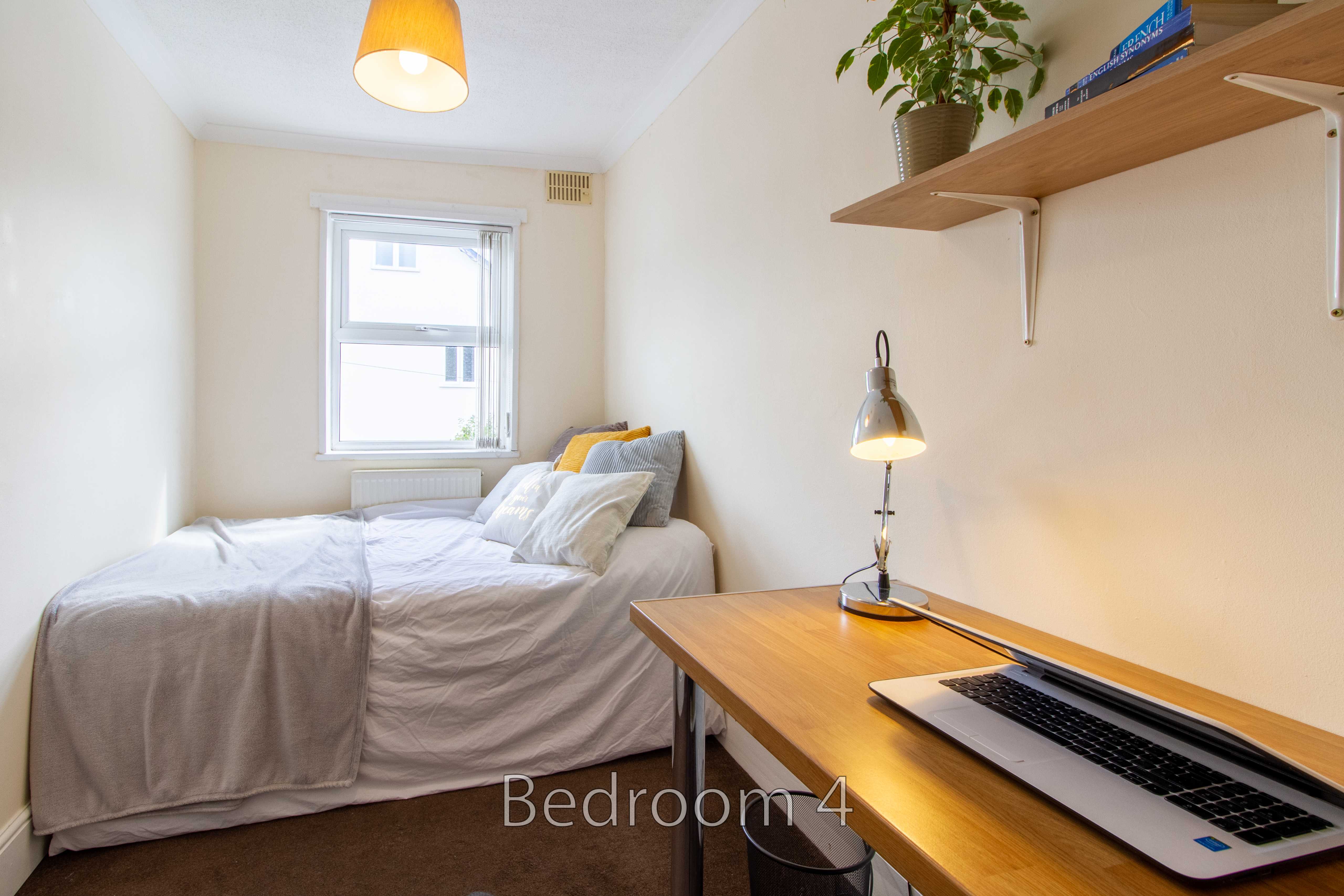 A compact bedroom with a single bed, desk, laptop, wall shelves, and a window.