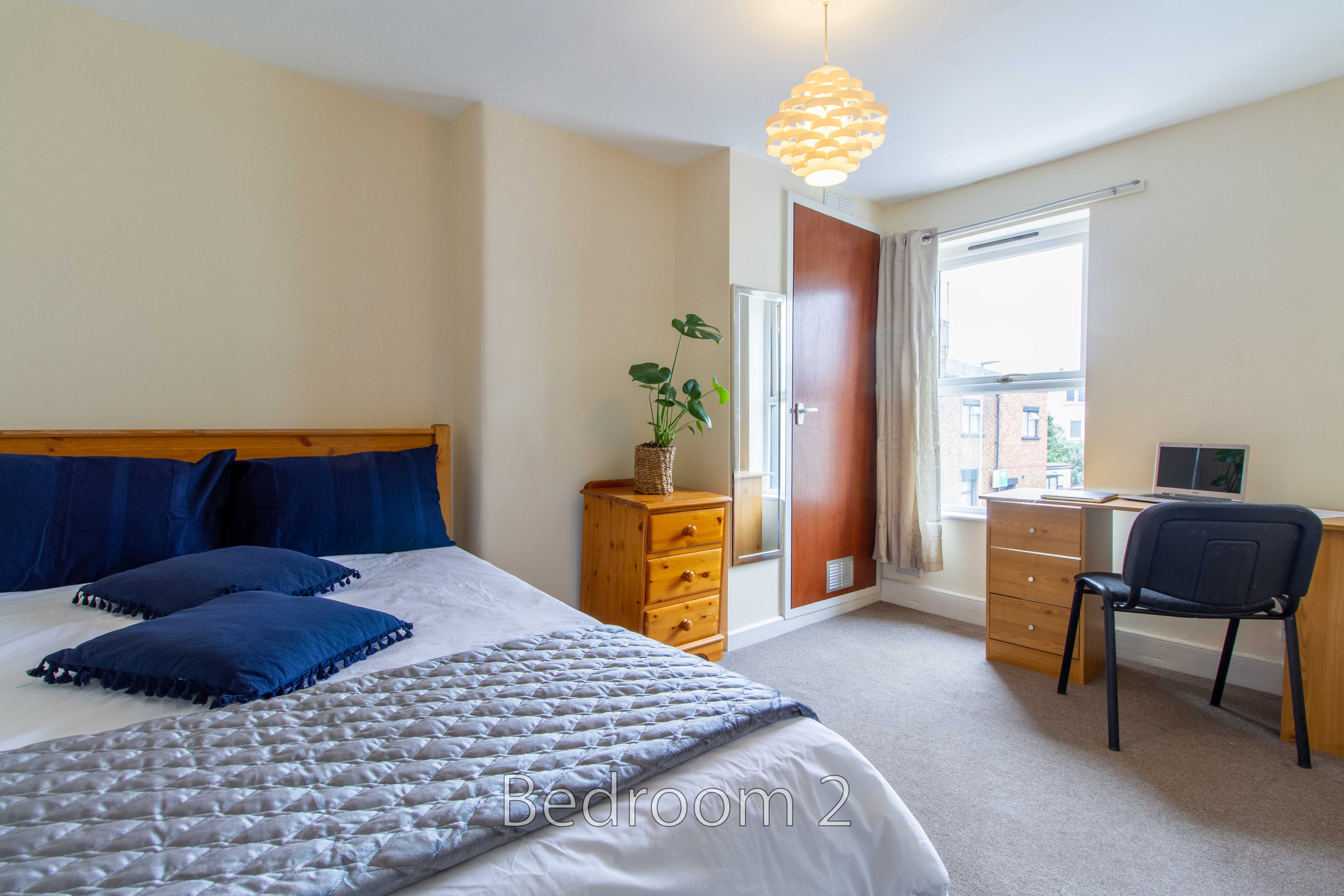 A bedroom with a double bed, bedside chest, desk, chair, window and open door.