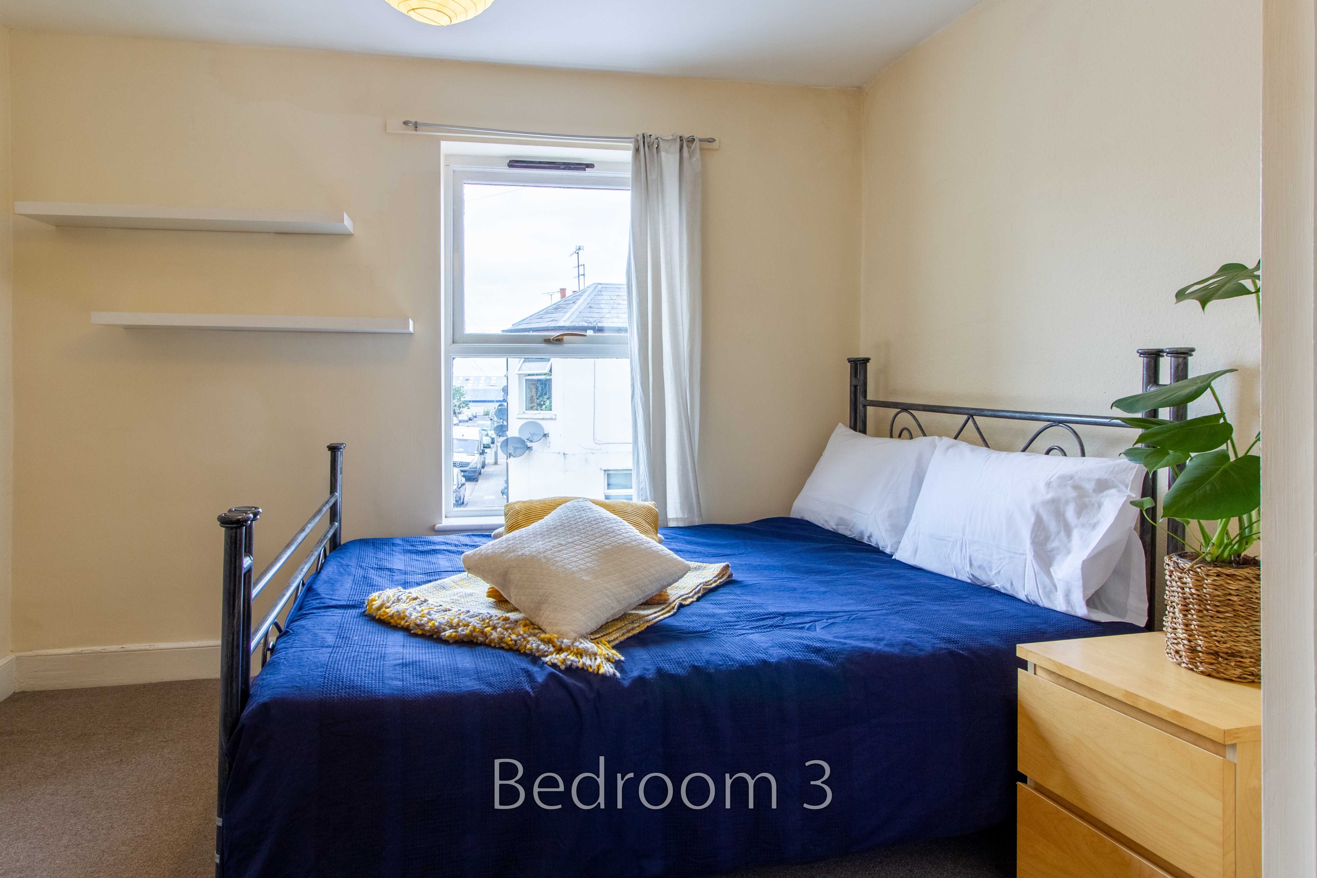 A bedroom with a blue bed, white pillows, wall shelves, a window and a chest of drawers.