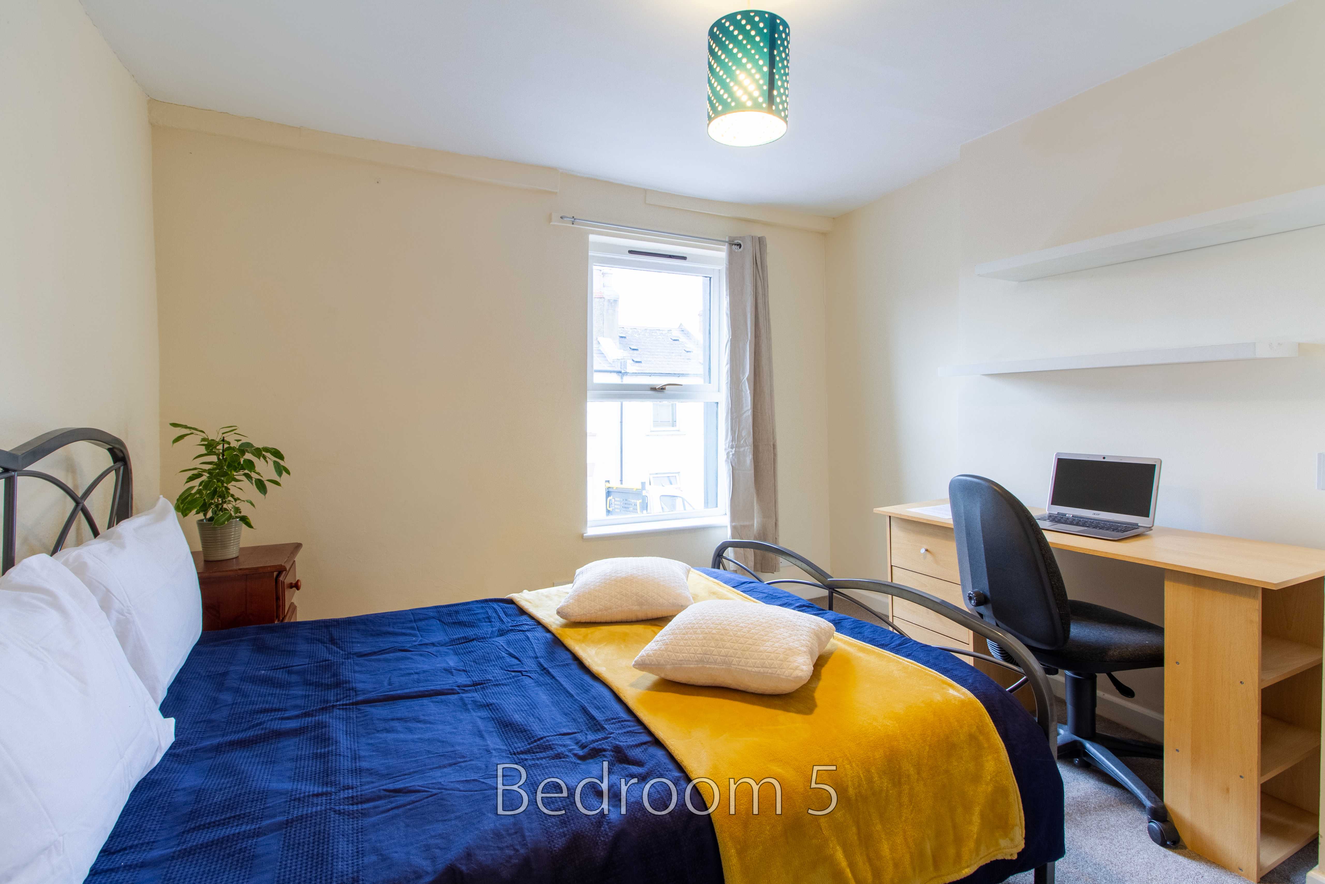 A bedroom with a bed, desk, chair, window and wall shelves.