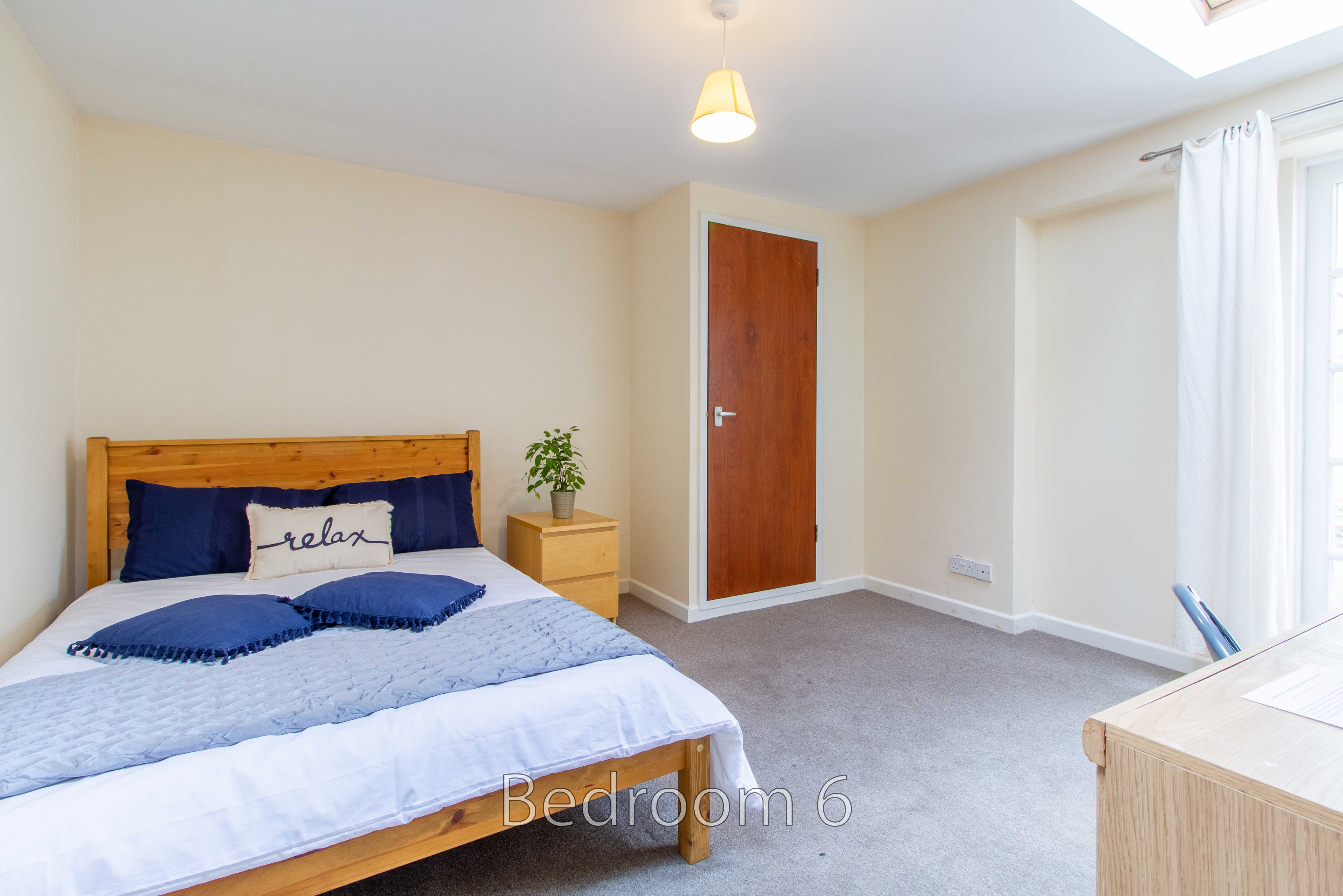 A bright bedroom with a double bed, bedside table, carpeted floor, door and window.