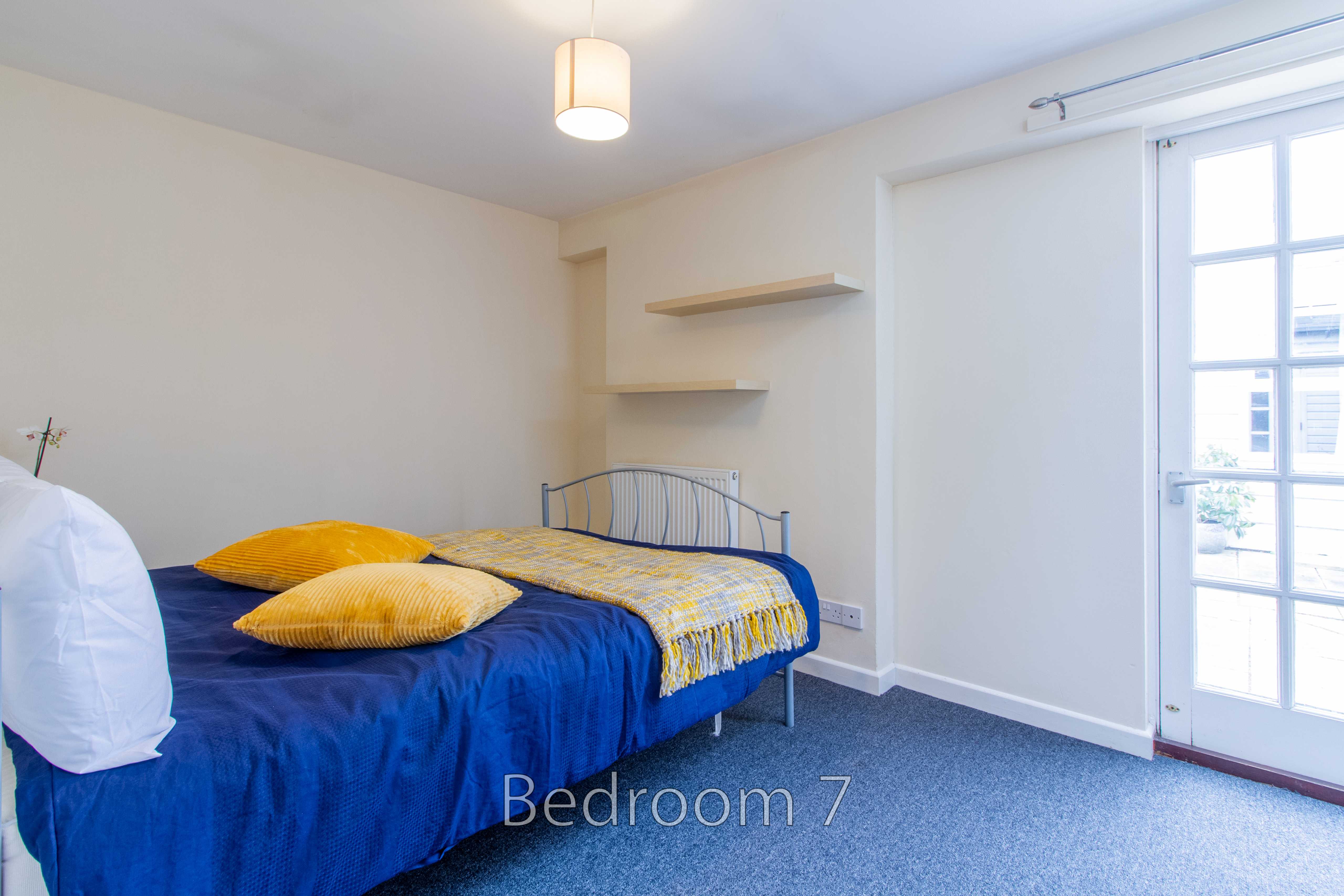 A bedroom with a metal bed, blue and yellow bedding, wall shelves, carpeted floor and a glazed door.
