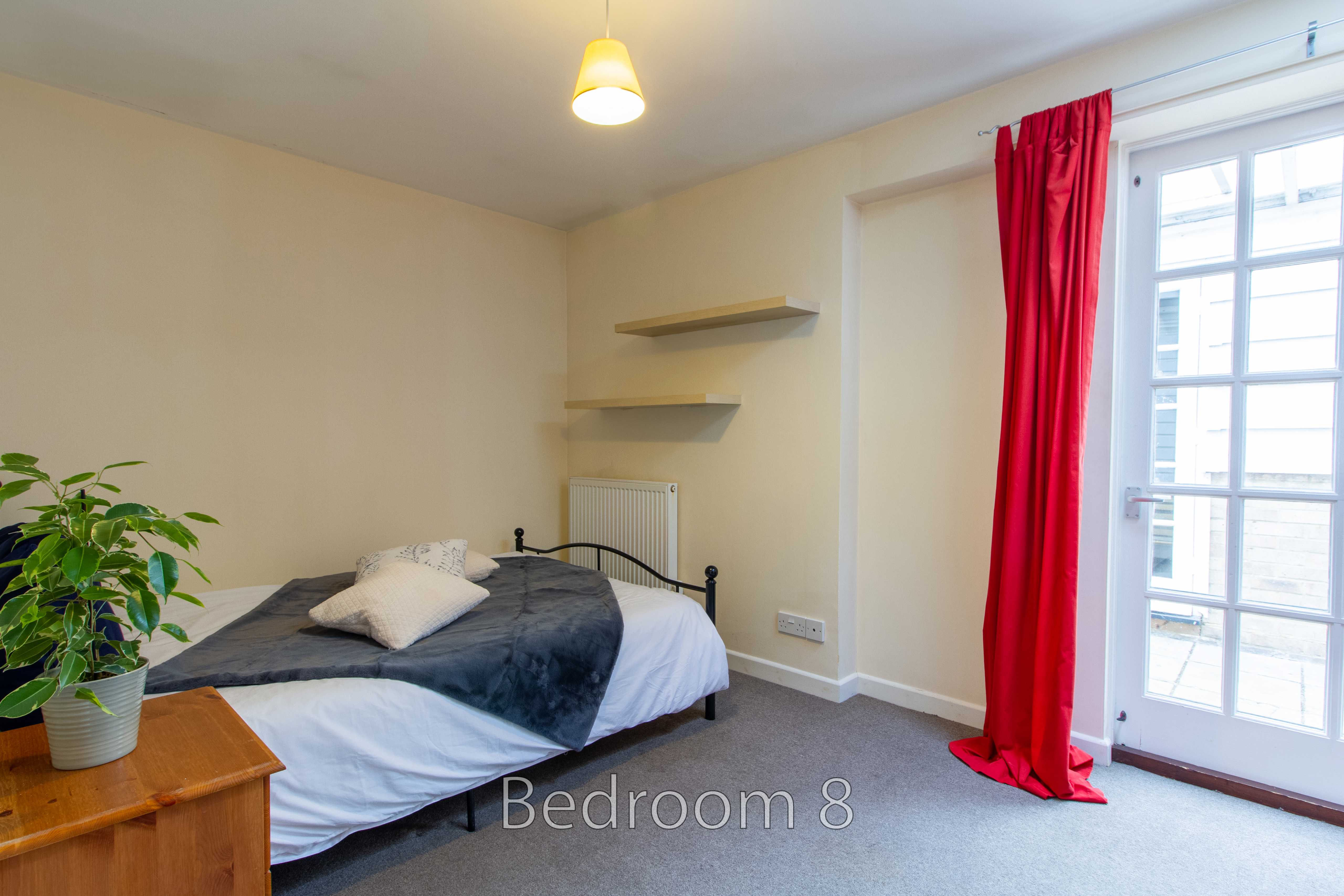 A bedroom with a bed, bedside table, wall shelves, radiator, plant, and glass-paned door with red curtains.