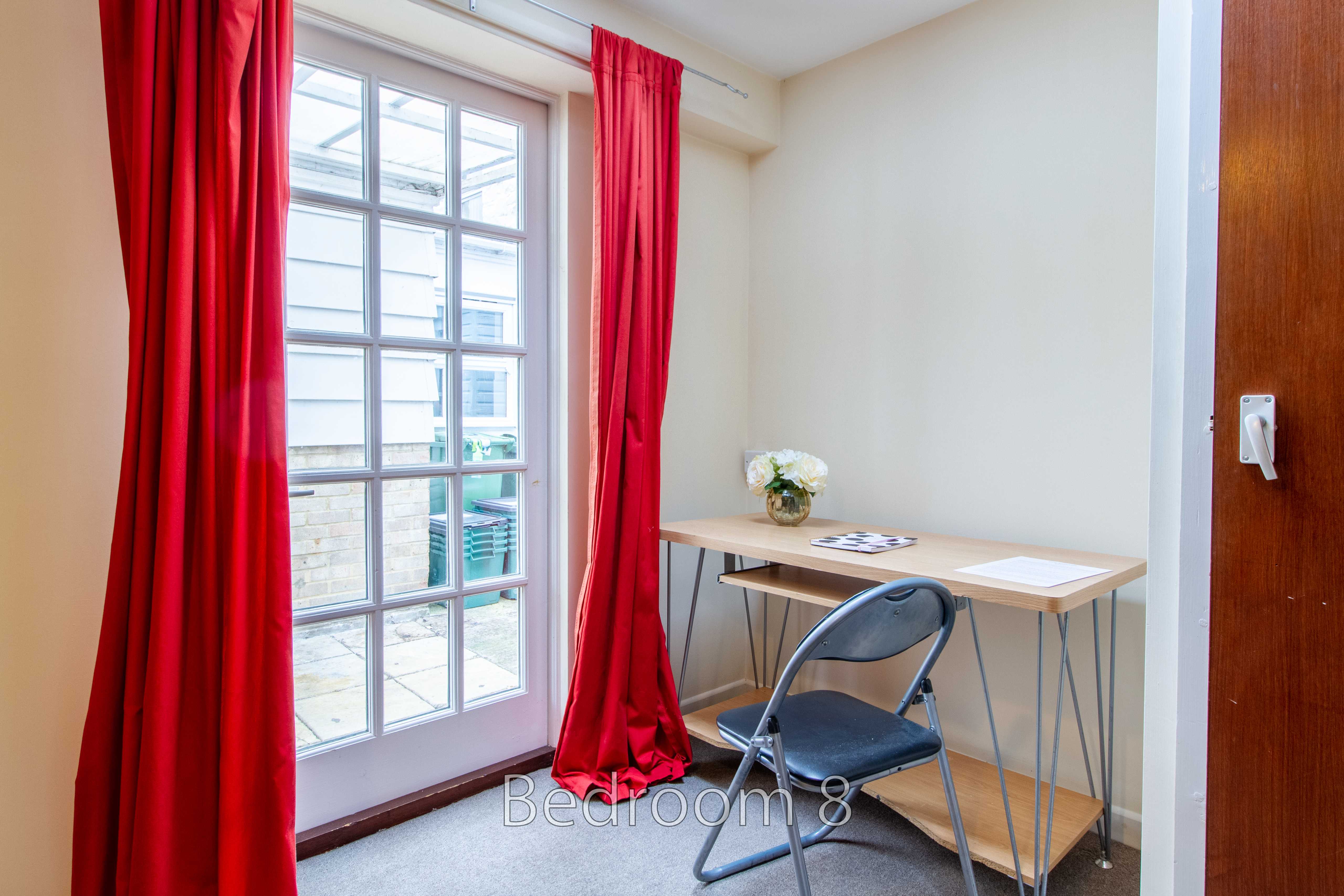 A small bedroom with a tall window, red curtains, a desk, and a folding chair.