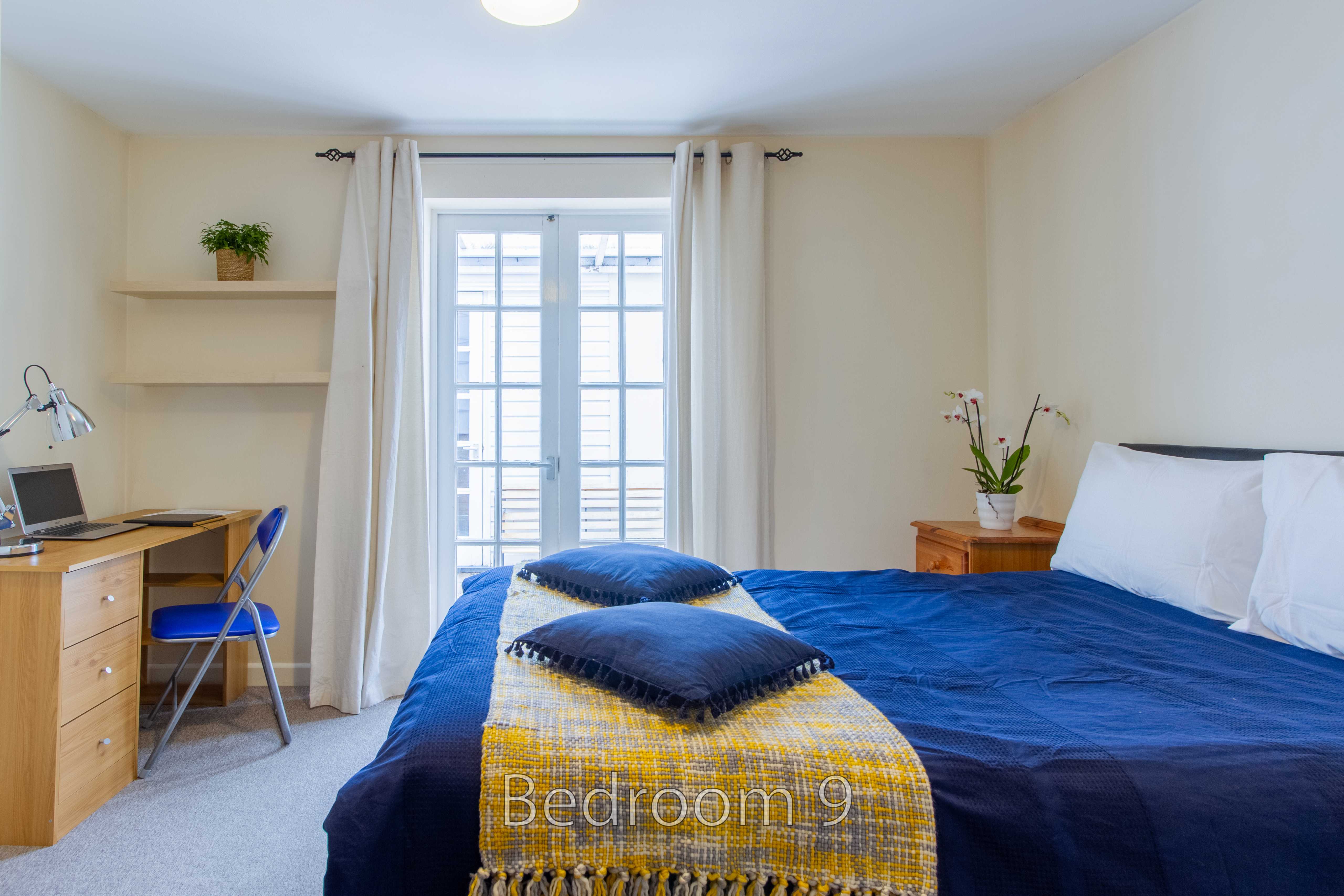 A furnished bedroom with a double bed, blue bedding, desk, chair, shelves, and a window.