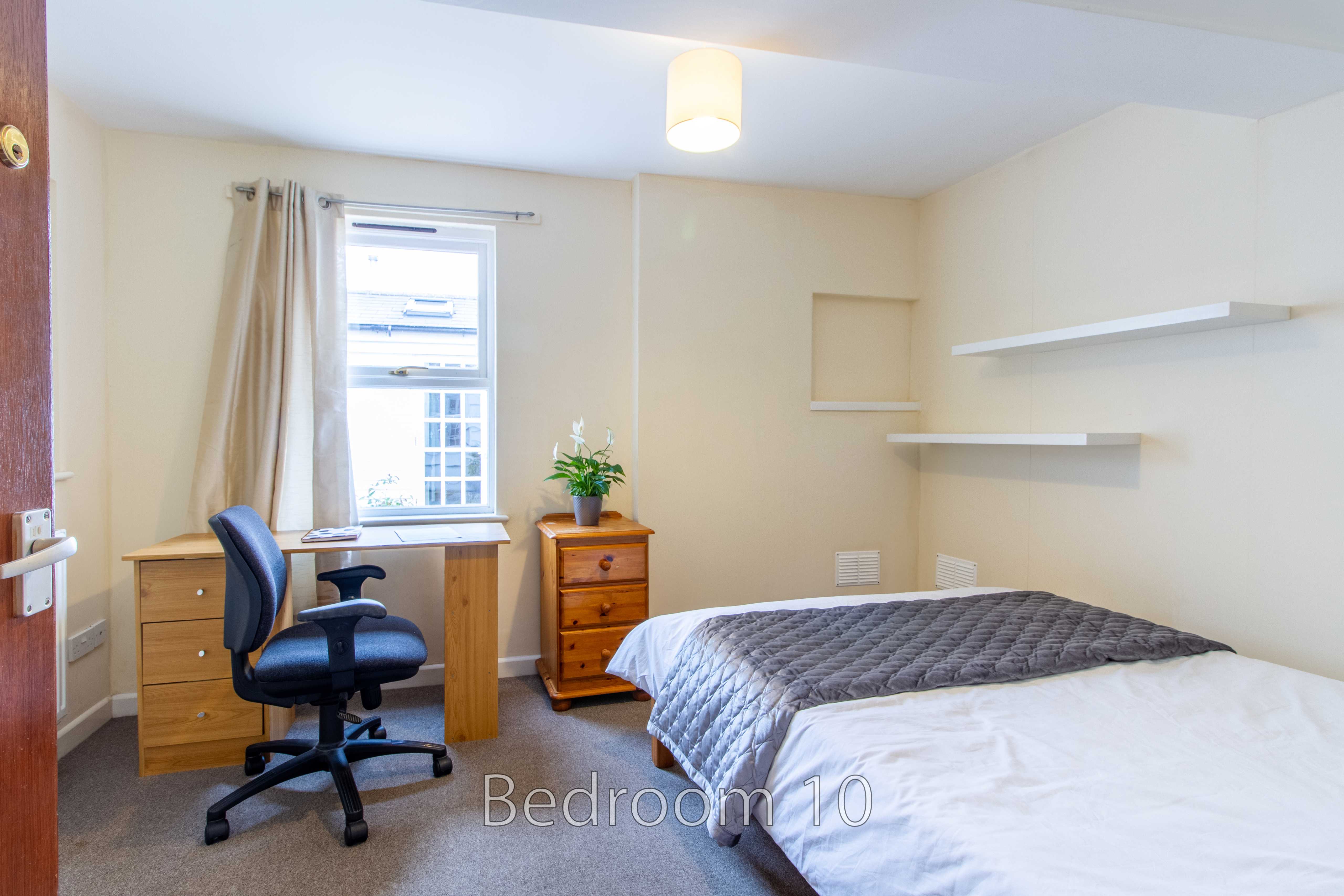 A bedroom with a double bed, desk, office chair, drawers, window and wall shelves.