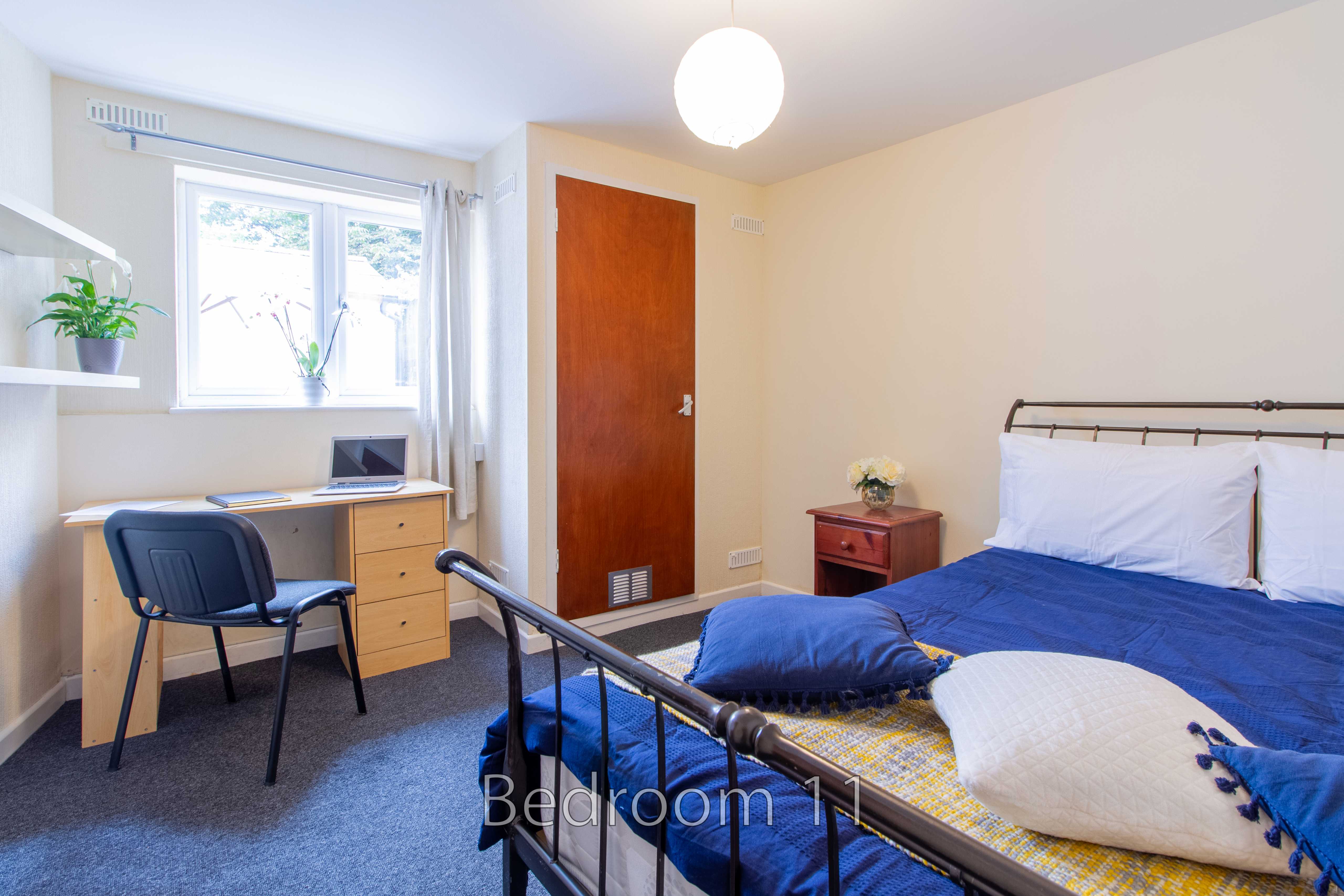 A bedroom with a double bed, desk, chair, bedside table, window and wooden door.