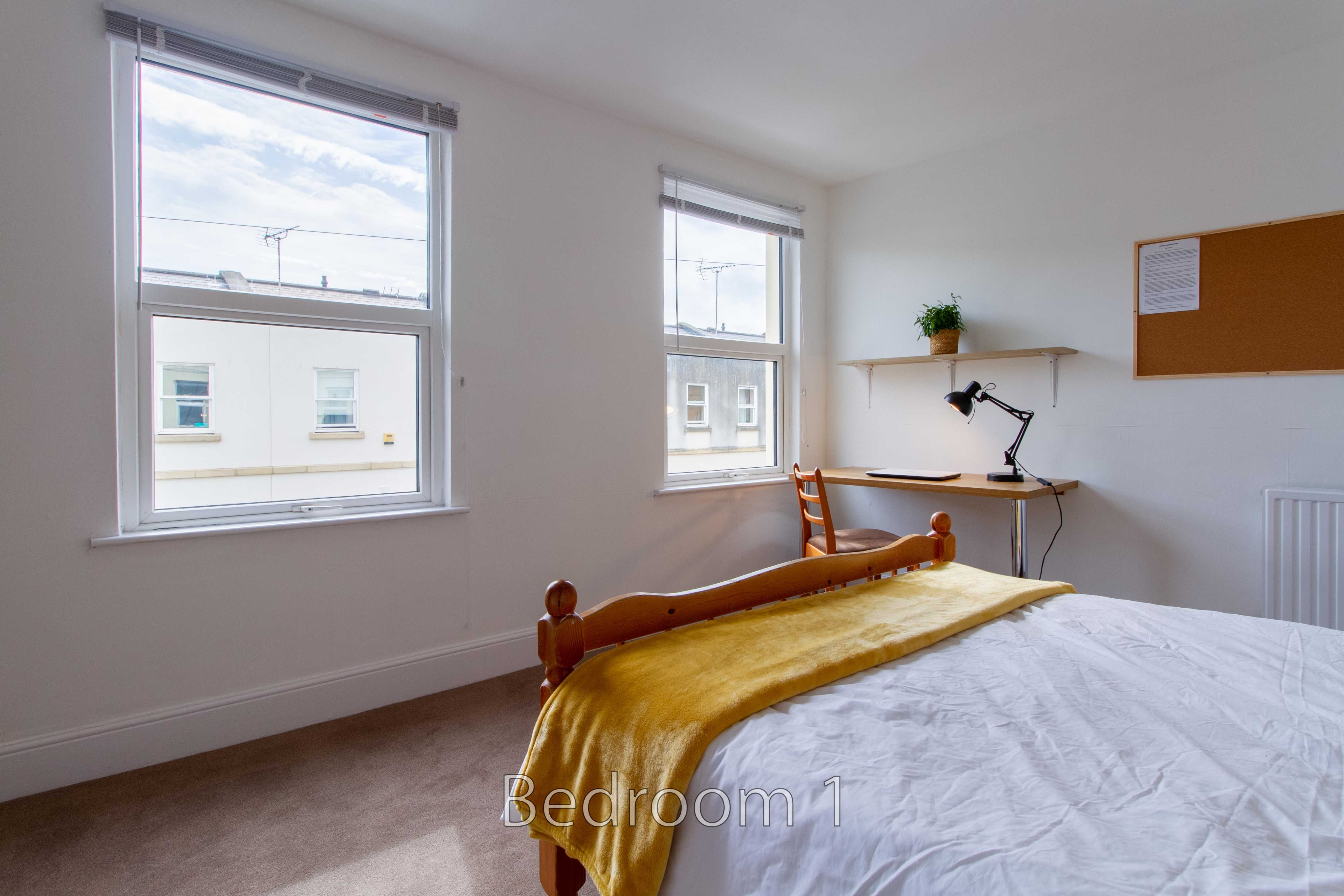 Bedroom with a double bed, two windows, a desk, shelving and a corkboard.