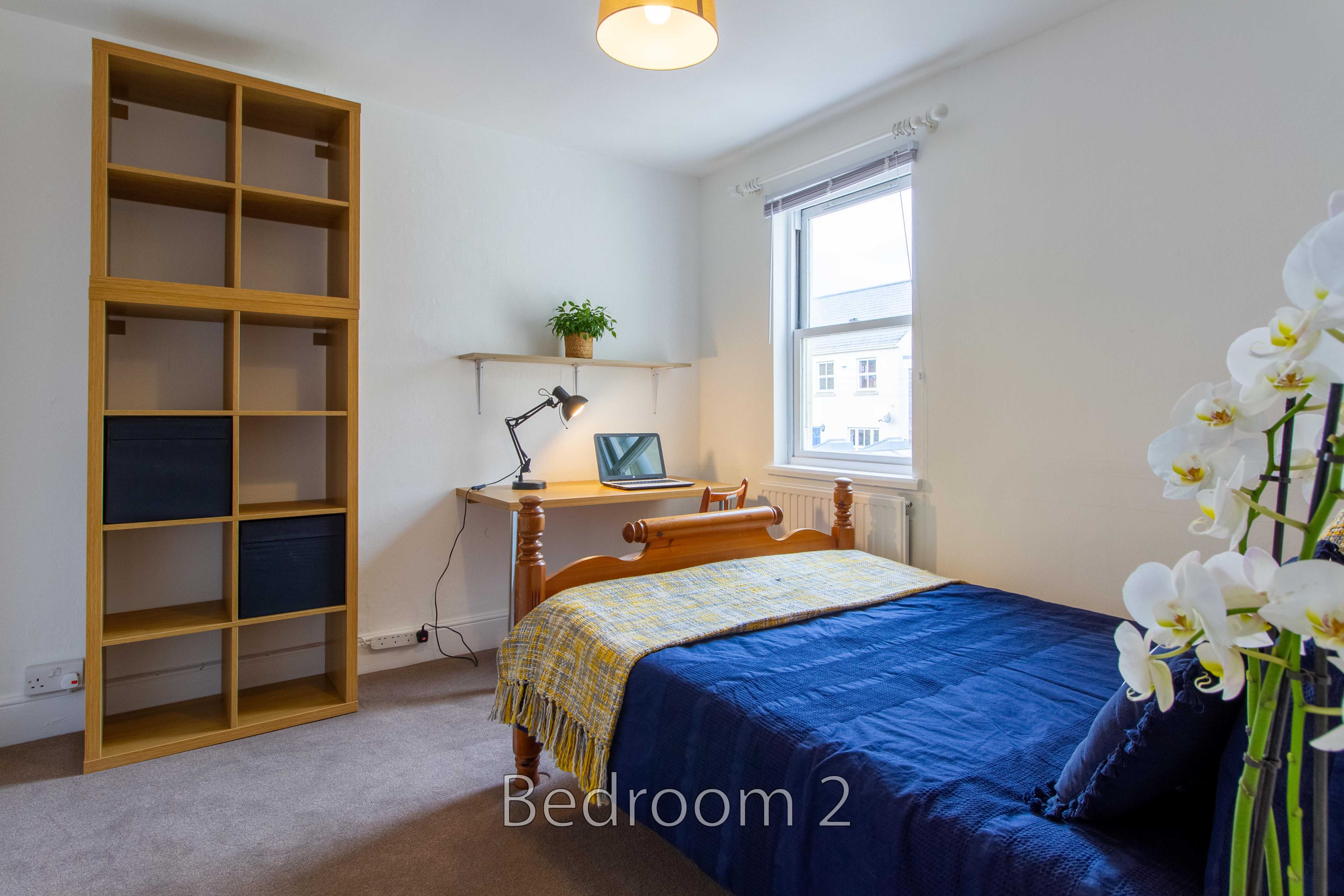 A furnished bedroom with a bed, desk, shelving, window and white orchid flowers.