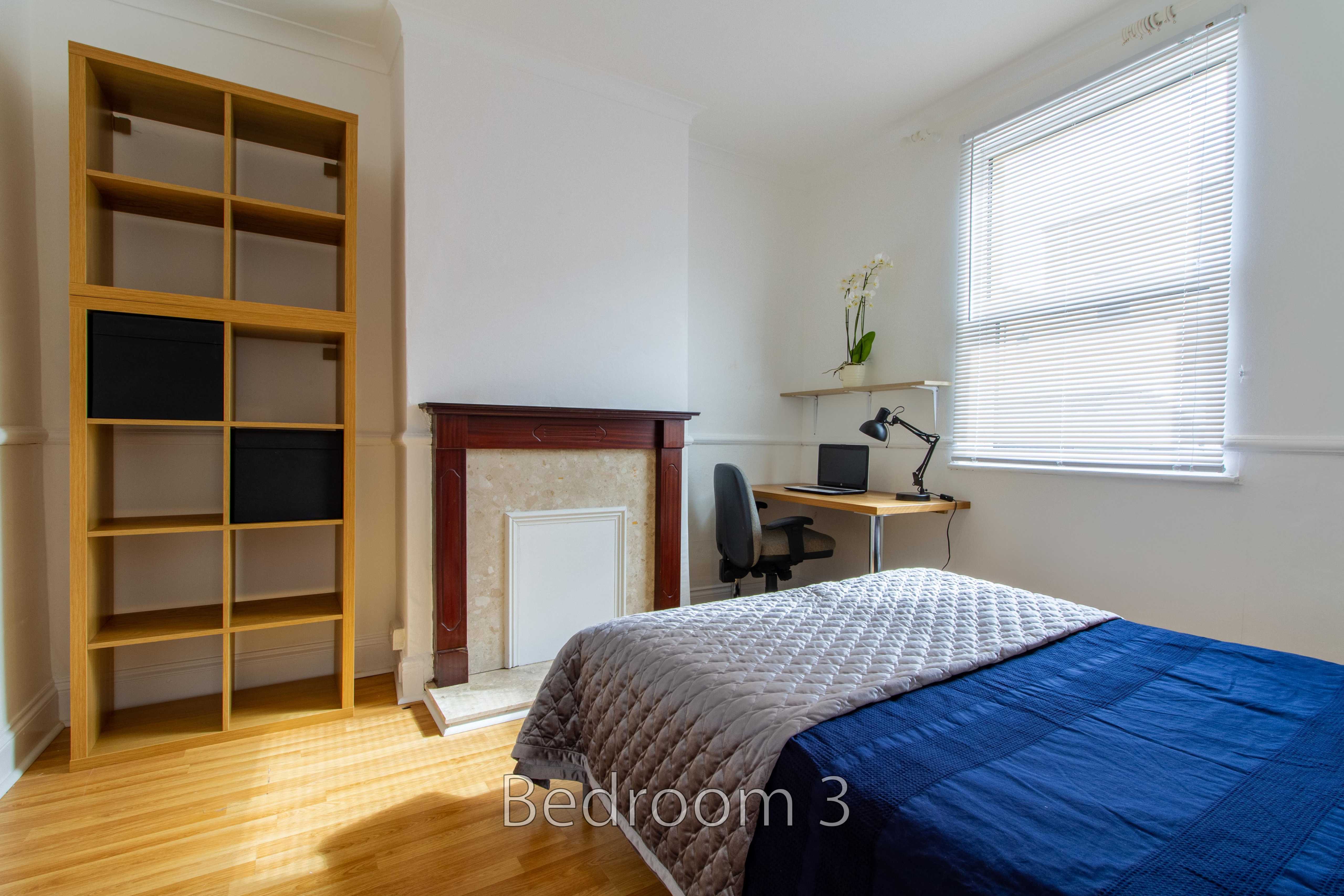 A bedroom with a bed, wooden shelving, fireplace, desk, chair and window blinds.
