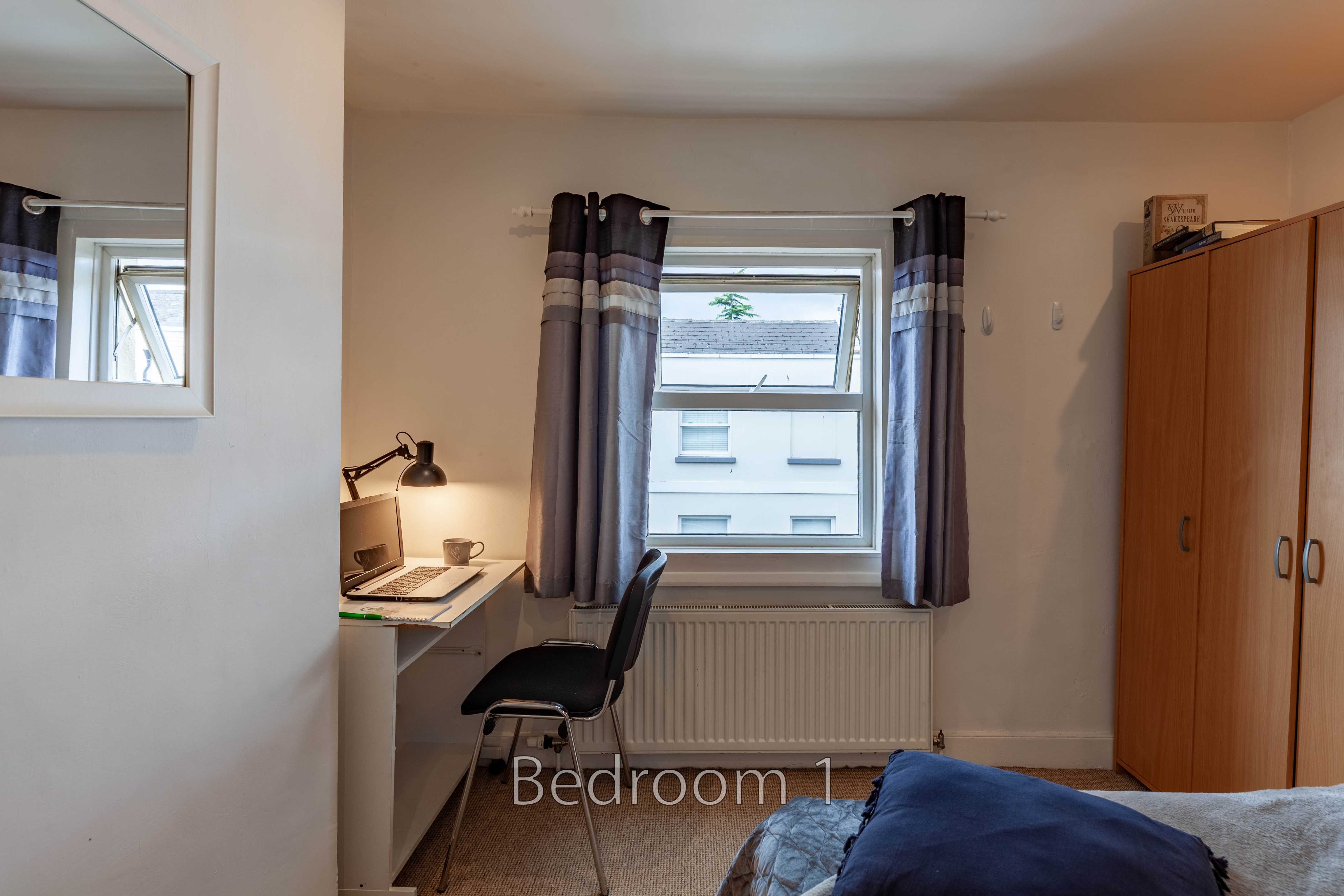 A bedroom with a bed, desk, chair, wardrobe, radiator and window.