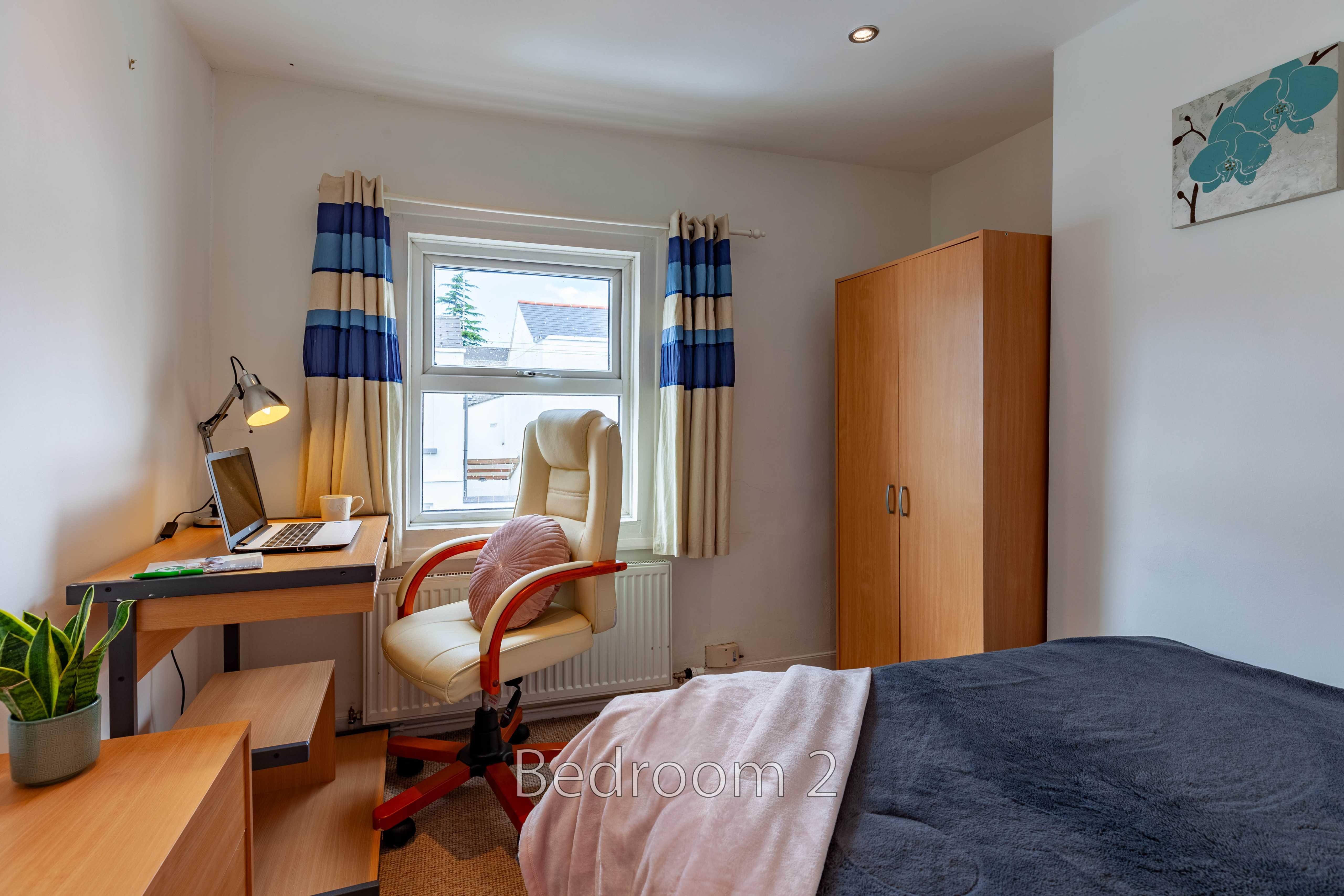 A furnished bedroom with a bed, desk, chair, wardrobe, window and blue-and-white curtains.