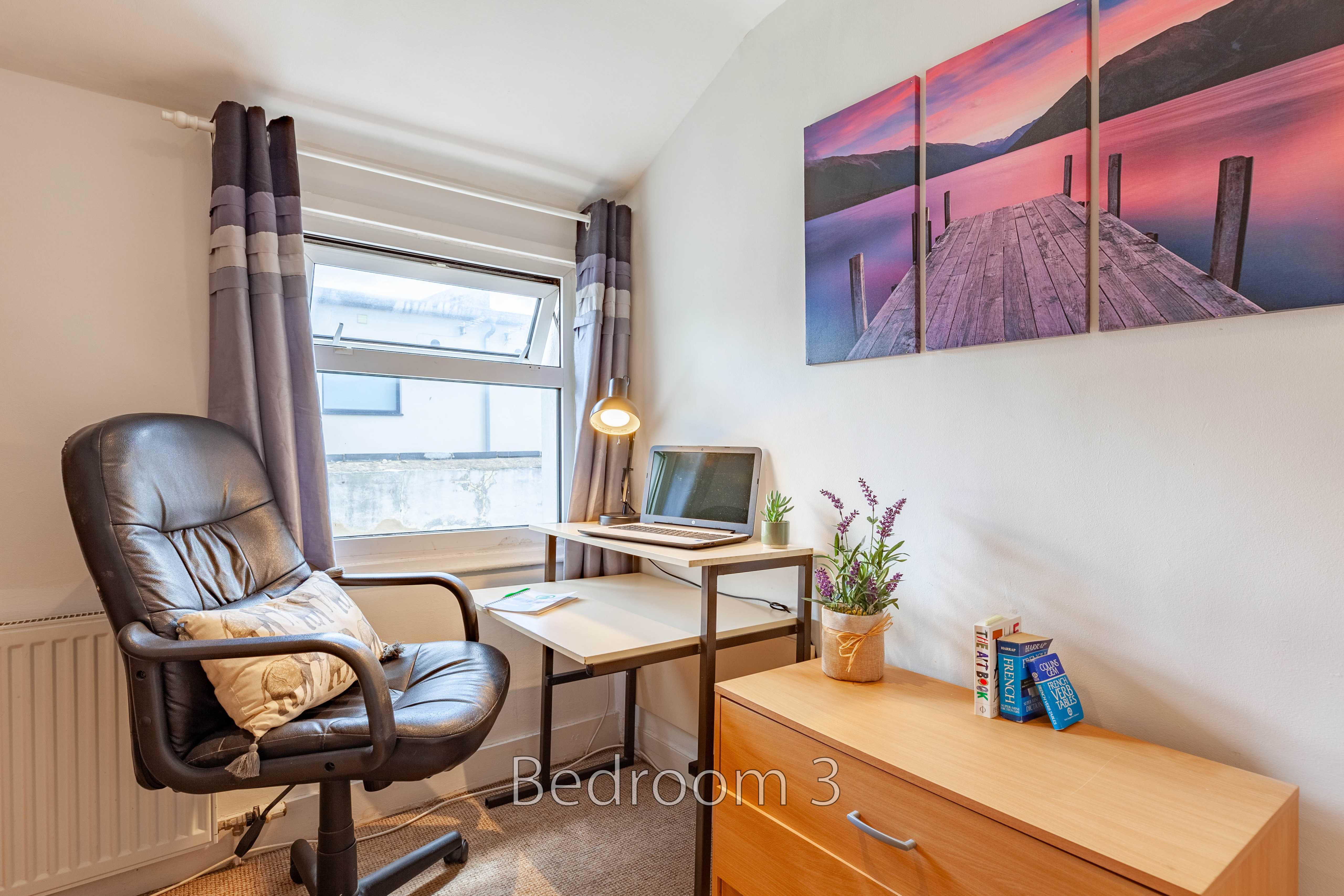 A furnished bedroom with a desk, office chair, chest of drawers, window and wall art.