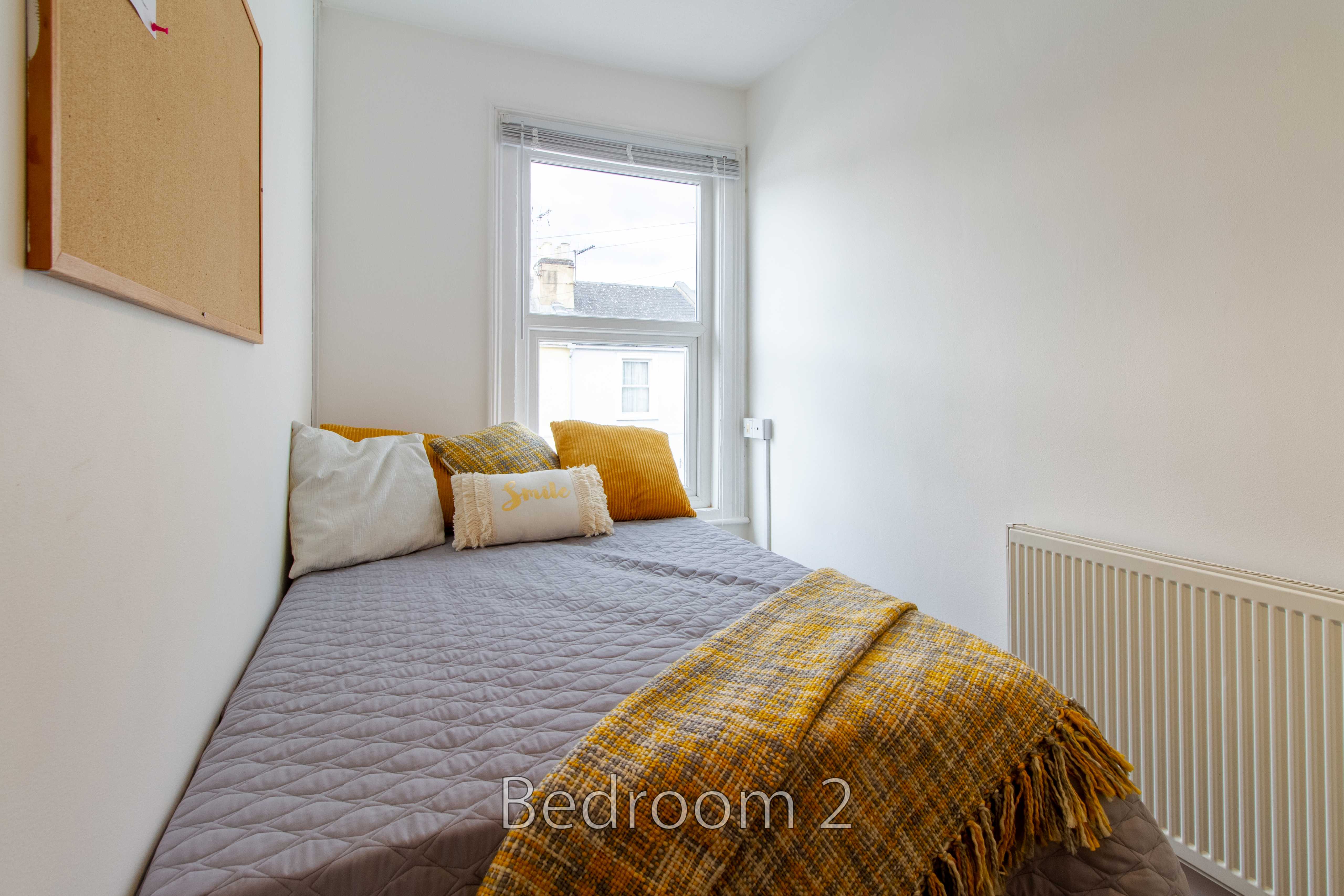 A small bedroom with a double bed, pillows, a mustard throw, a window and a radiator.