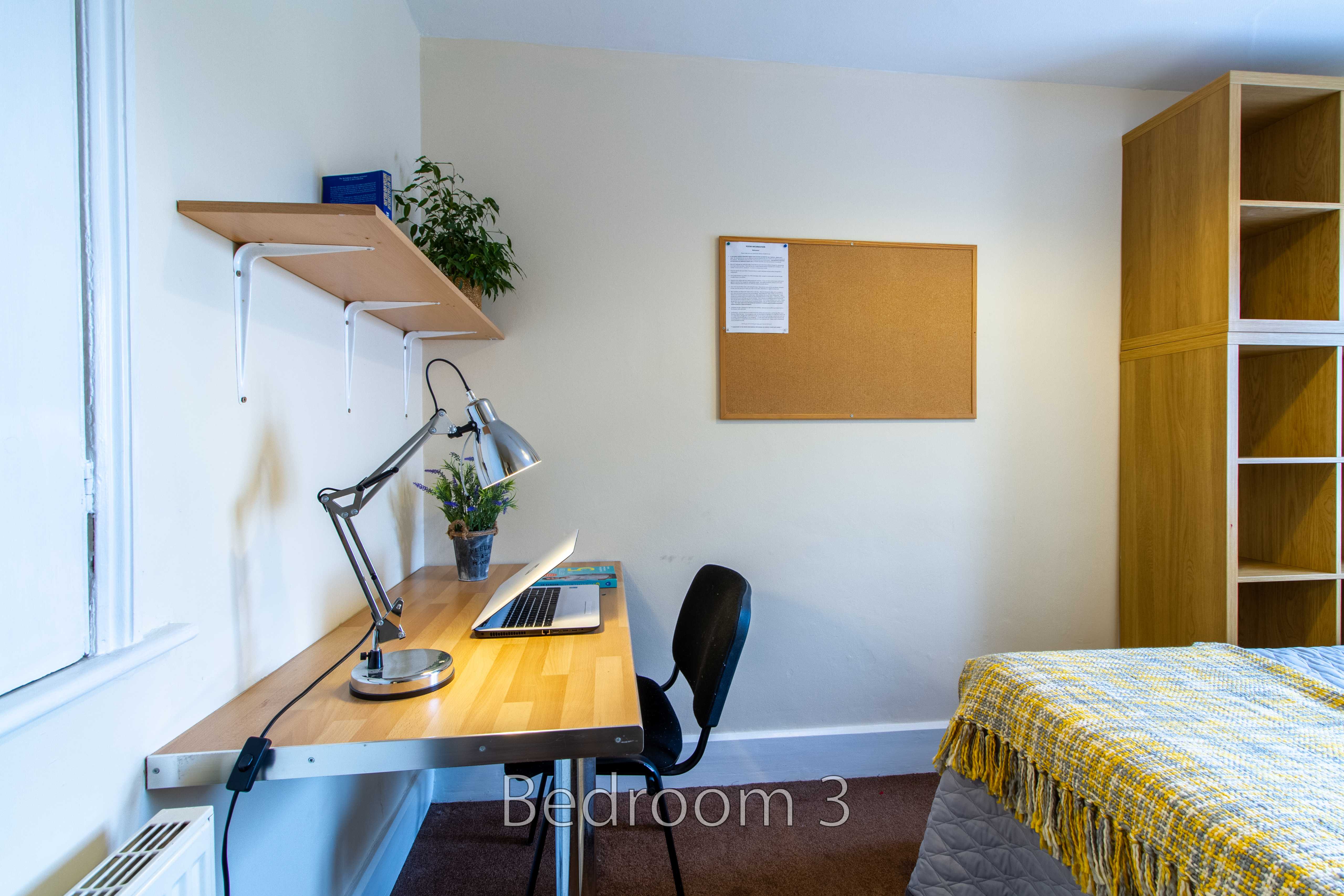 A bedroom with a desk, chair, shelving, bulletin board and single bed.