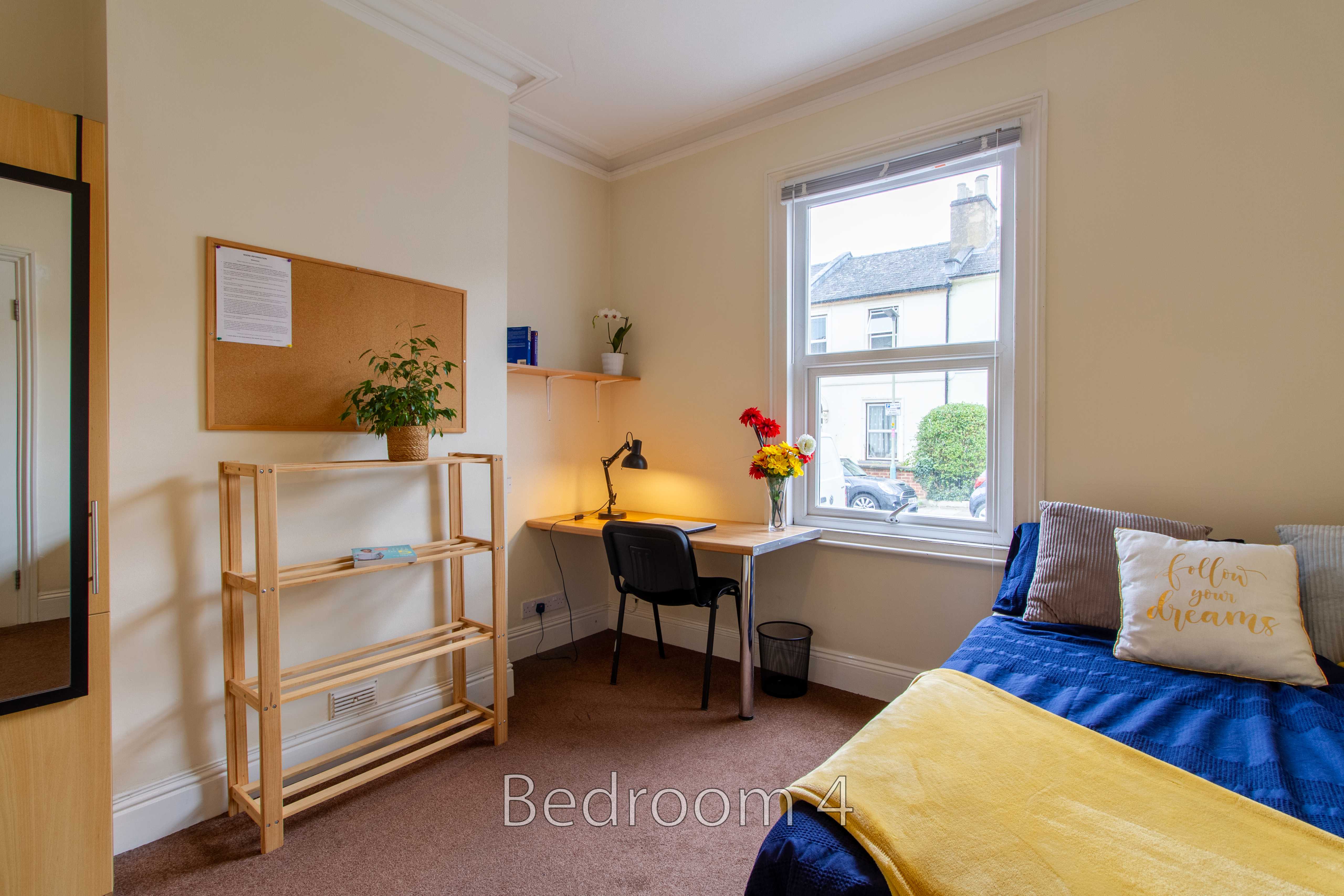 A furnished bedroom with a single bed, desk, chair, shelving unit and window.