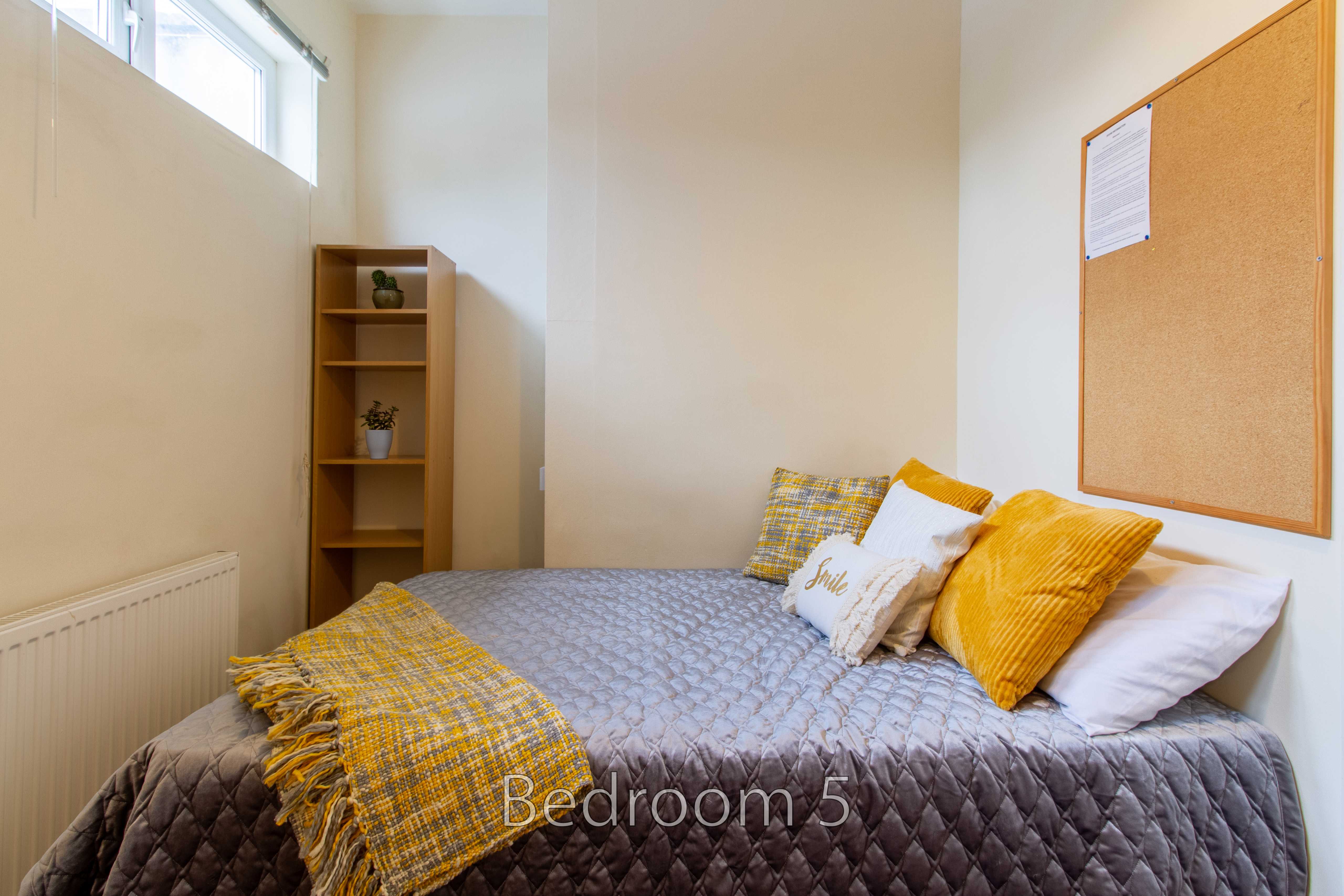 A bedroom with a bed, yellow and grey bedding, a wooden shelf, radiator and cork noticeboard.