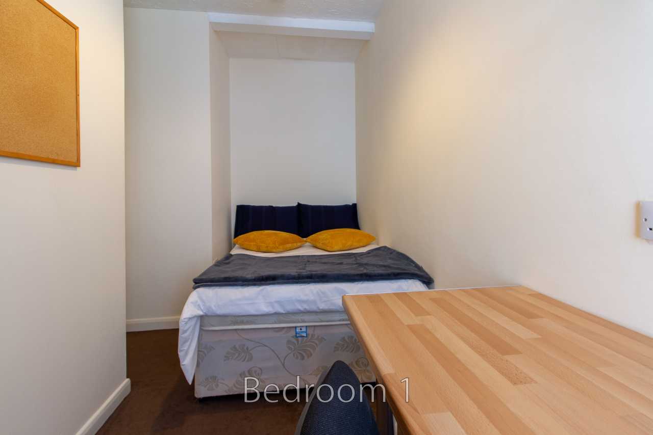 A small bedroom with a double bed, pillows, a wooden desk, and a corkboard on the wall.