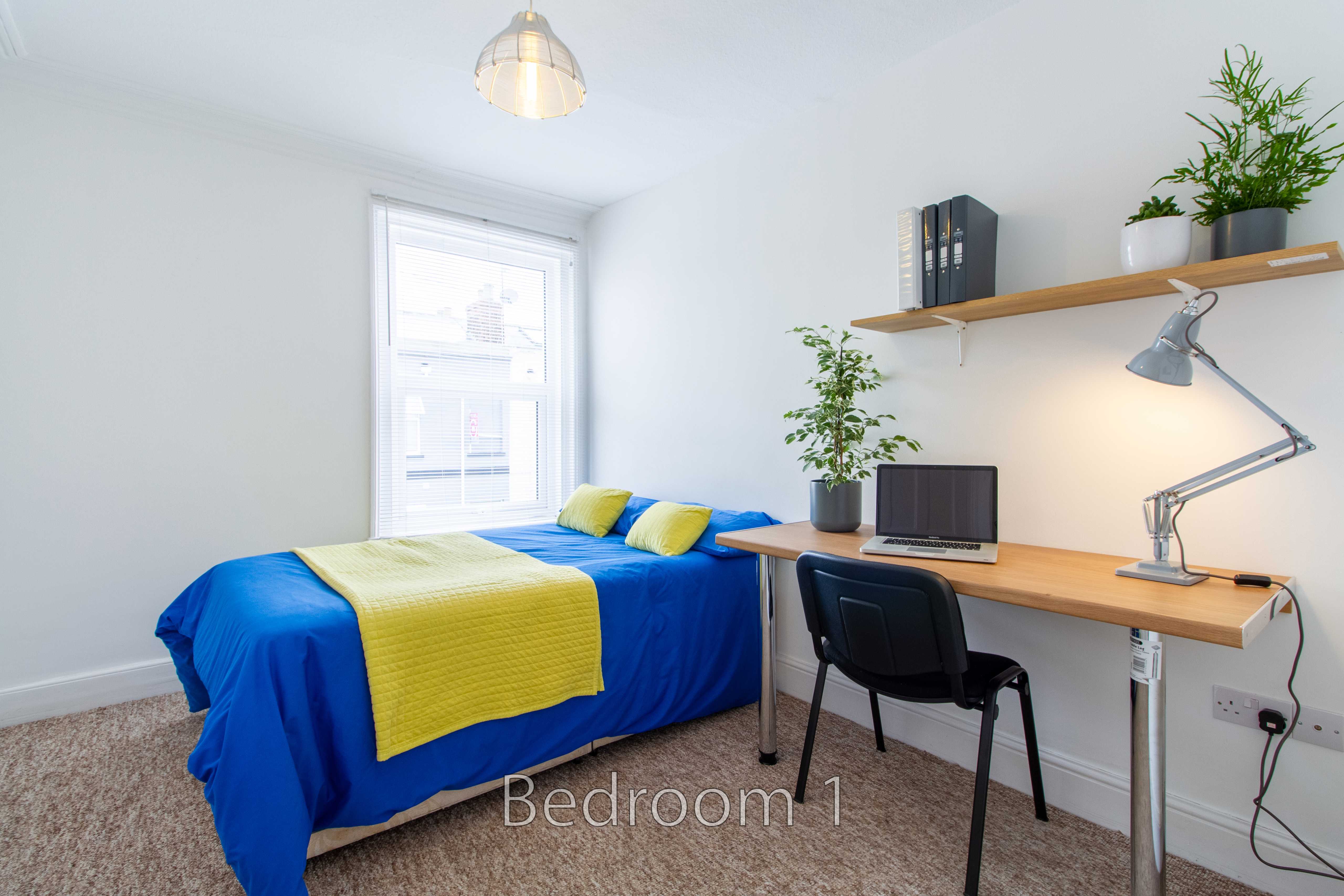 A furnished bedroom with a single bed, desk, chair, shelves and potted plants.