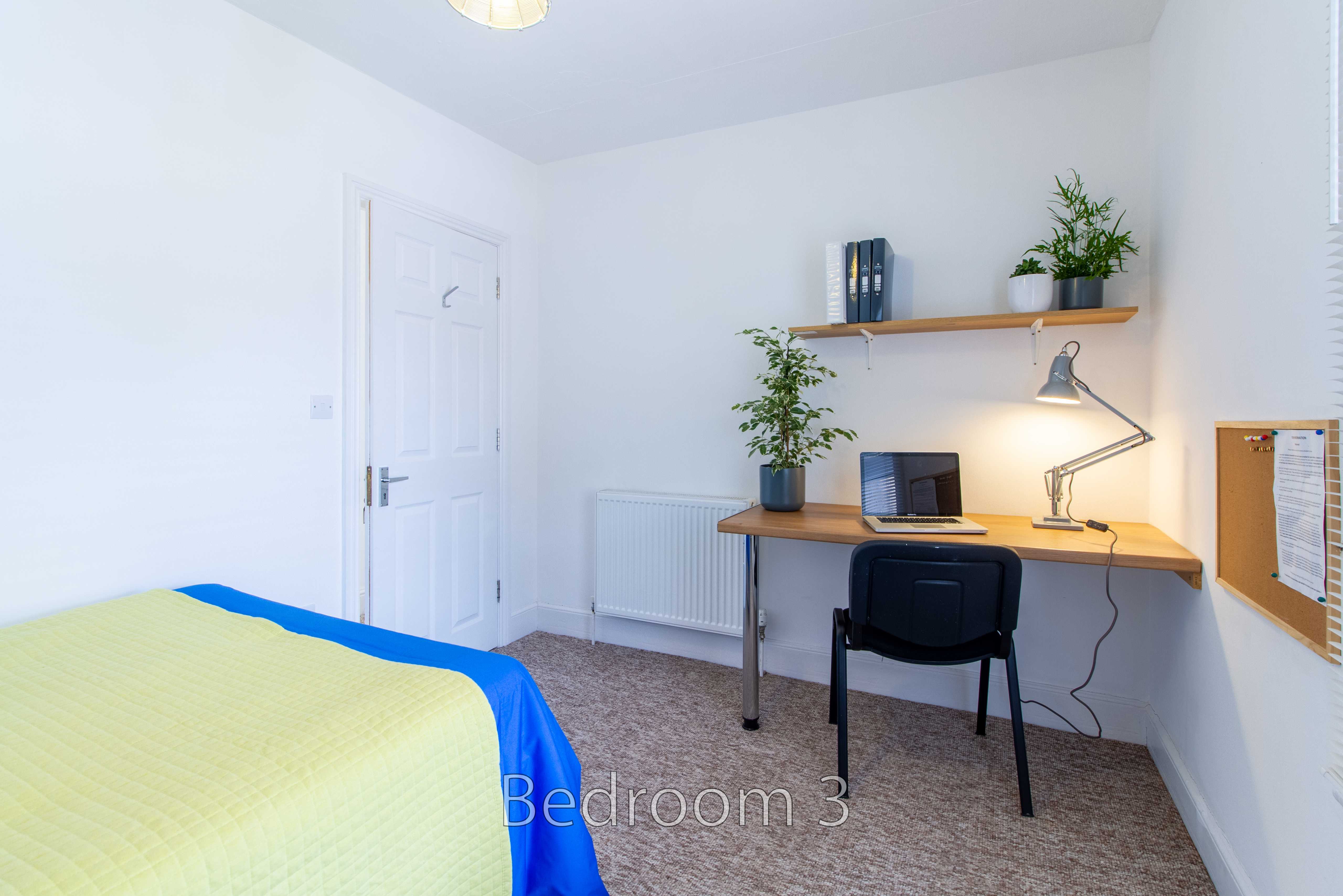 A bedroom with a single bed, desk, chair, shelves, radiator and wall noticeboard.