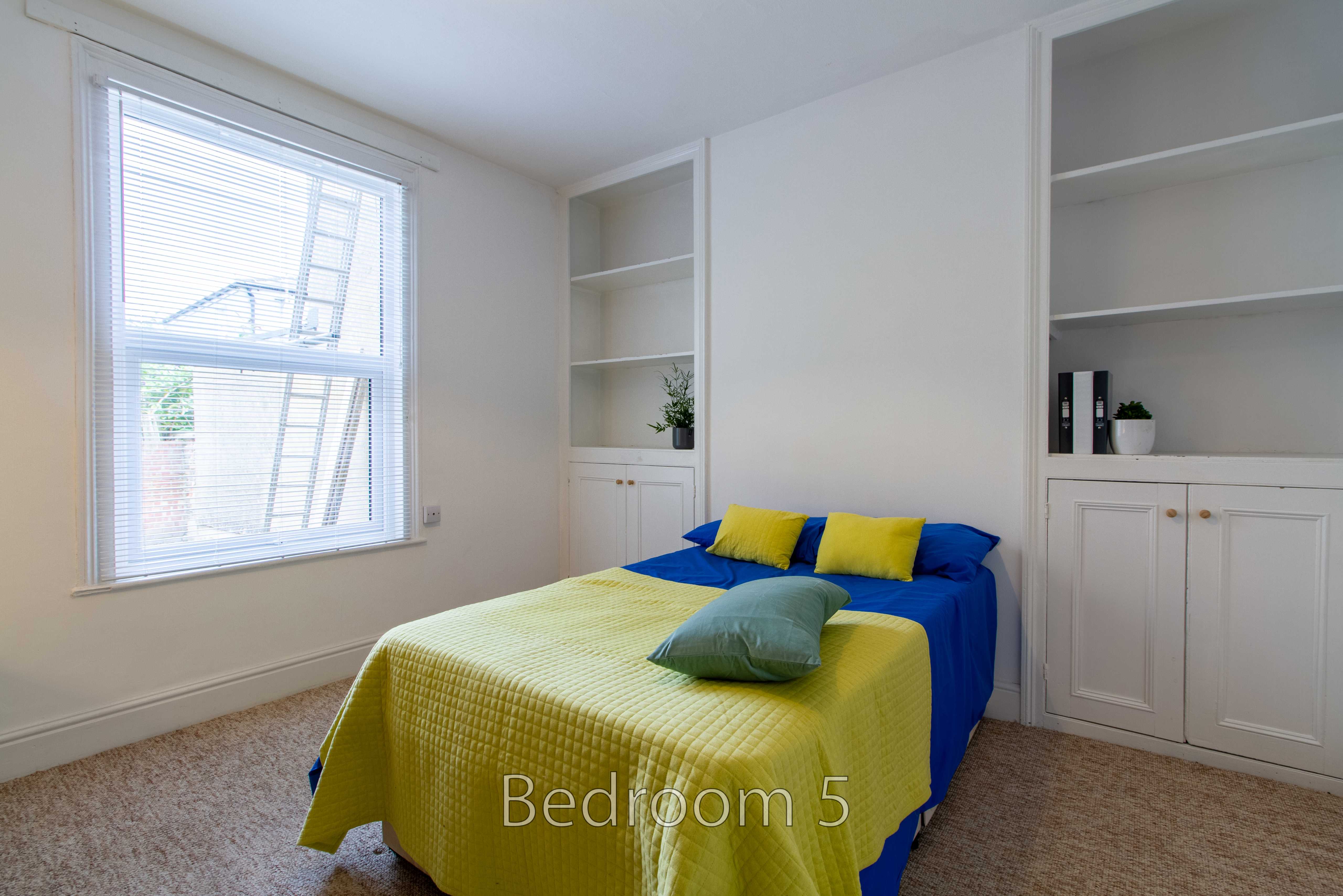 A bedroom with a bed covered by a bright yellow and blue throw, built-in shelving, a window, and beige carpet.