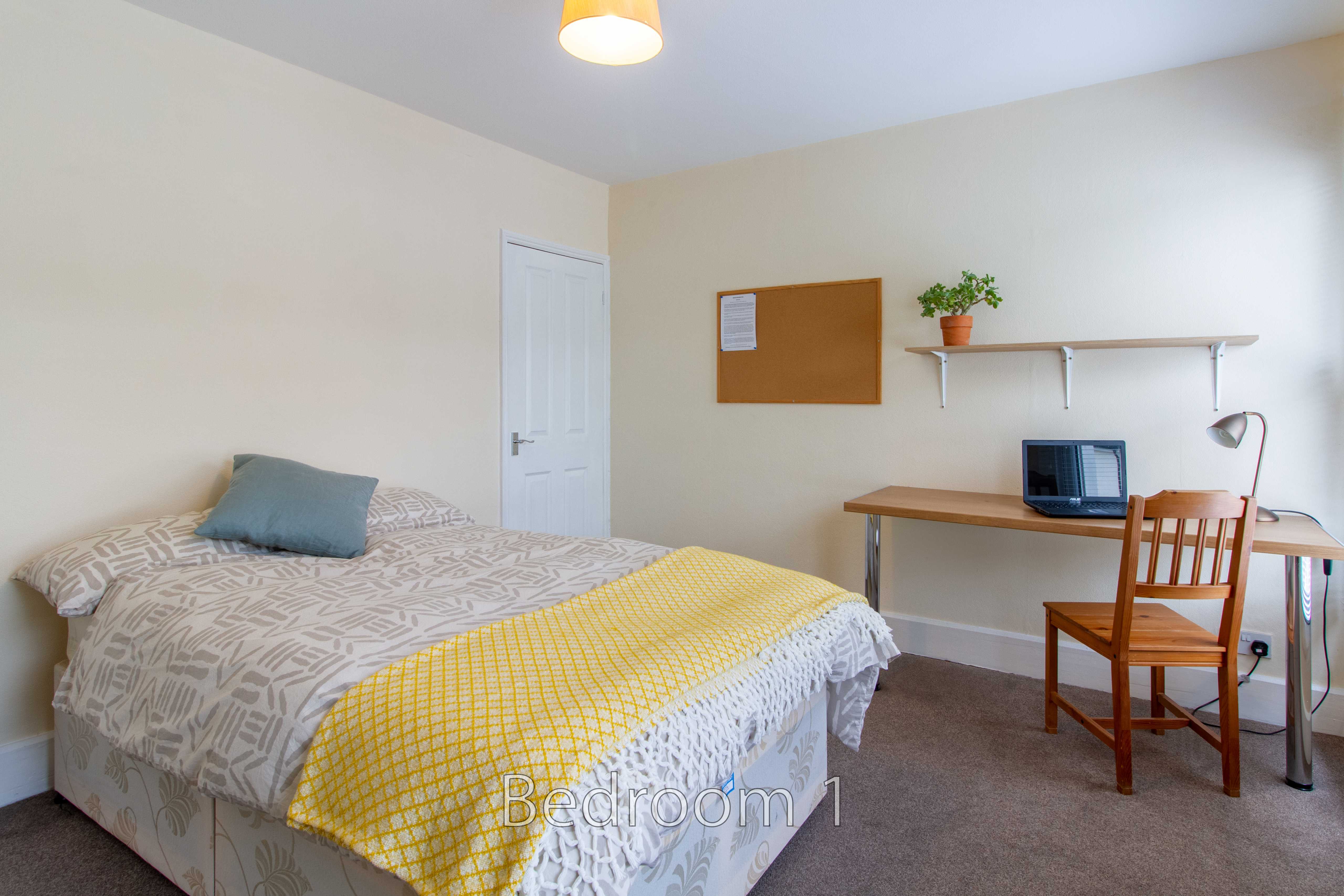 A furnished bedroom with a double bed, desk, chair, shelves and a door.