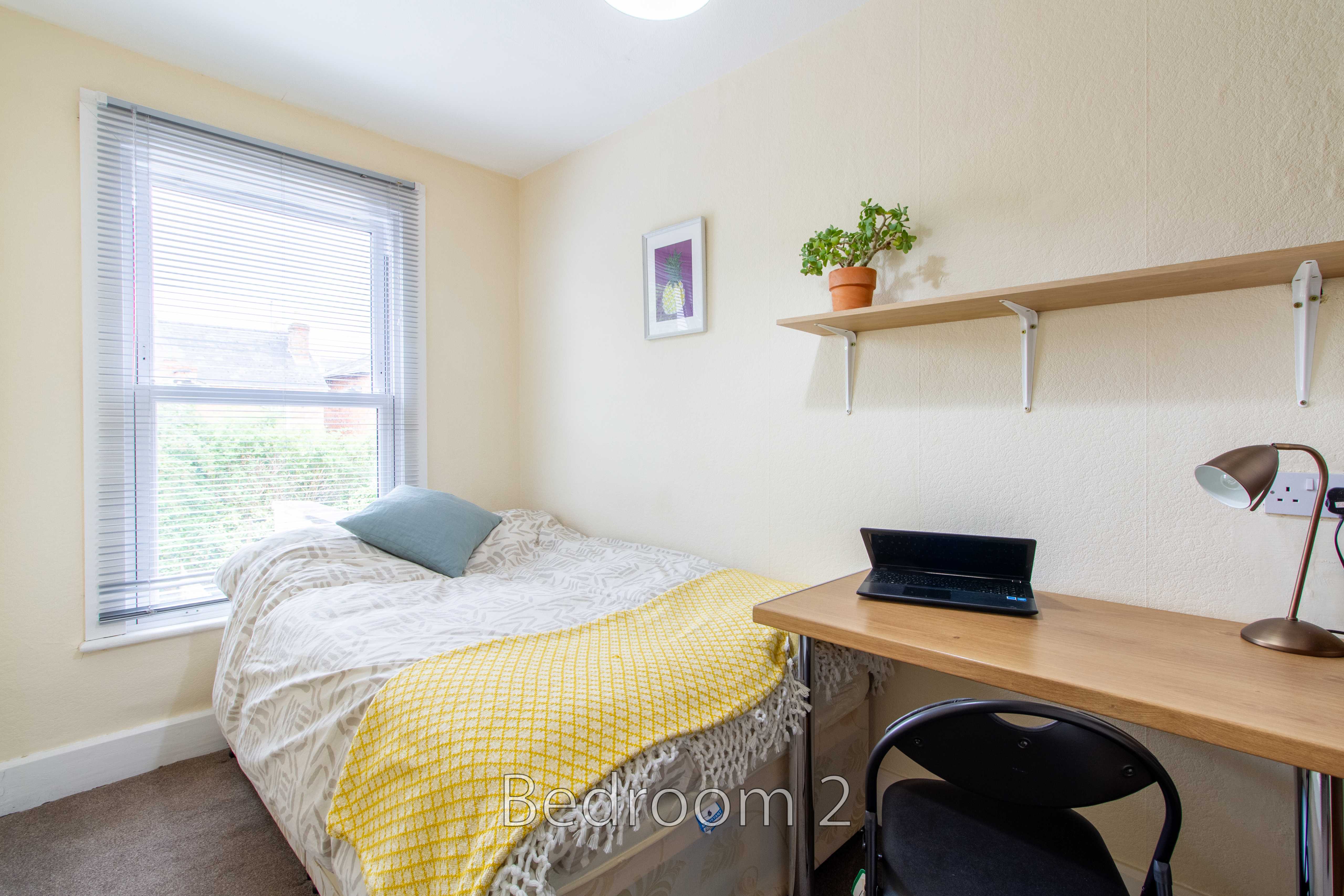 A bedroom with a single bed, desk and chair, window with blinds, shelf, and desk lamp.