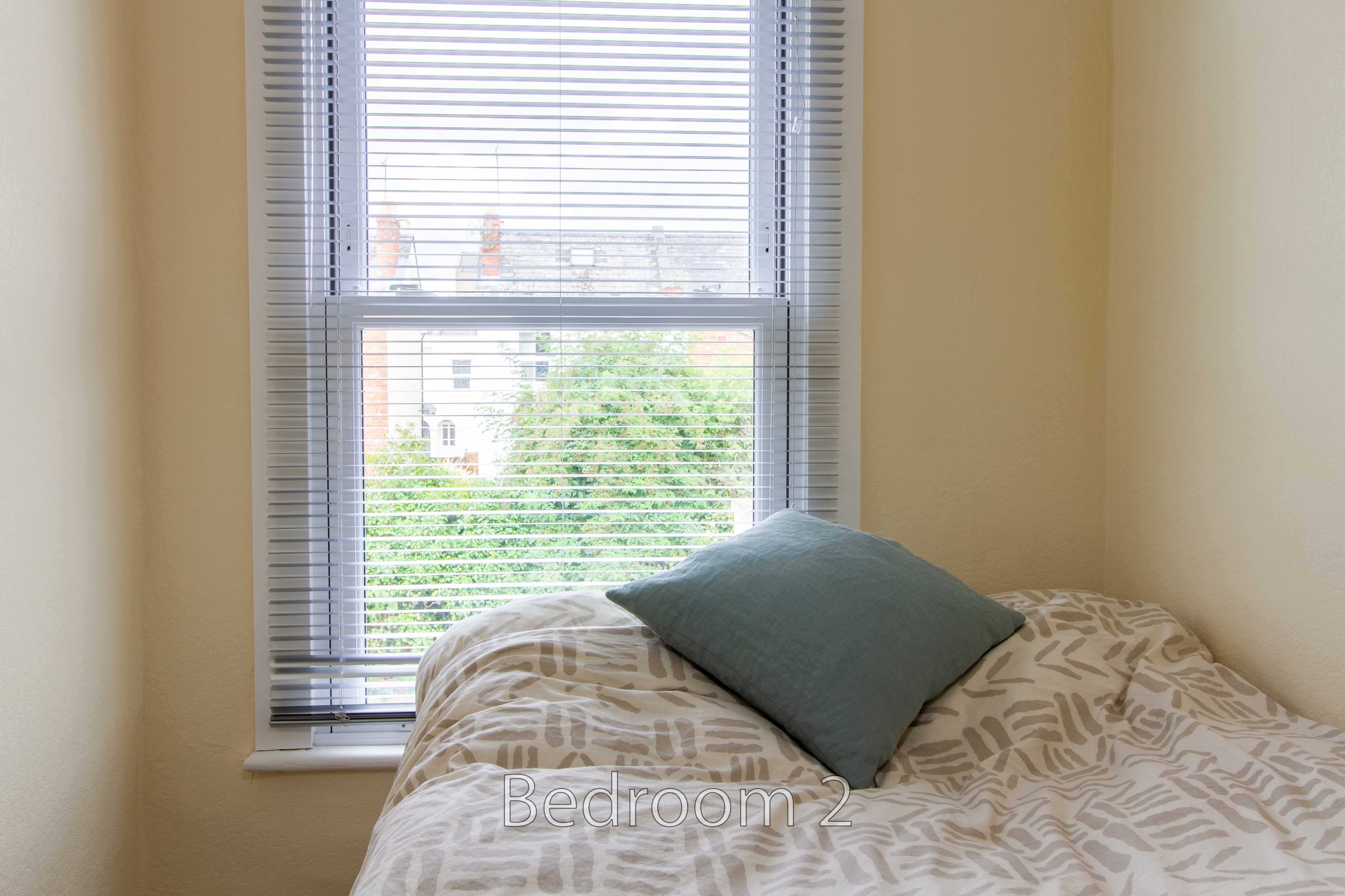 A small bedroom with a single bed, patterned duvet and blue pillow beside a window with blinds.