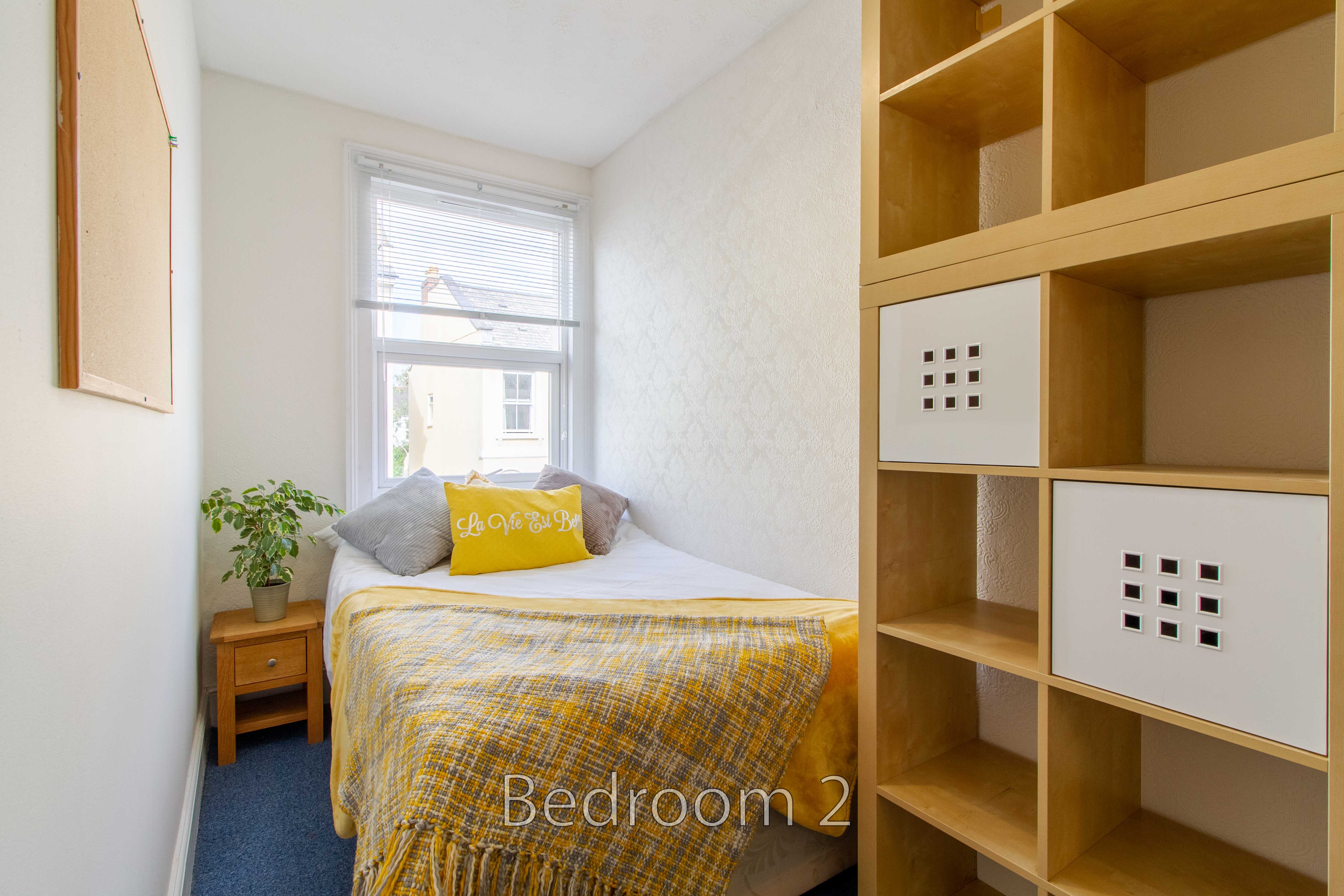 A small bedroom with a single bed, window, bedside table, plant and wooden shelving with storage boxes.