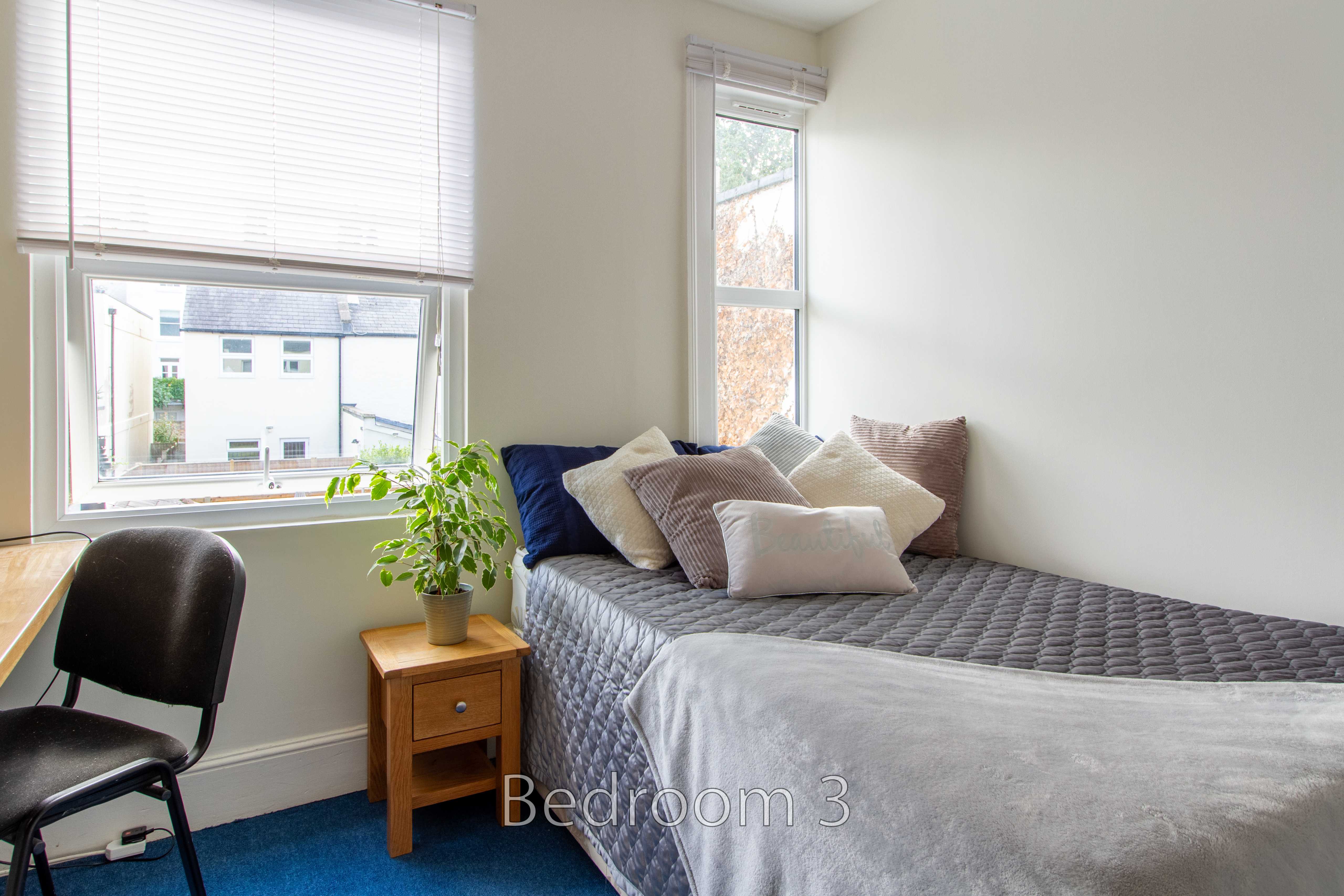 A bright bedroom with a single bed, pillows, windows with blinds, a bedside table and a chair.
