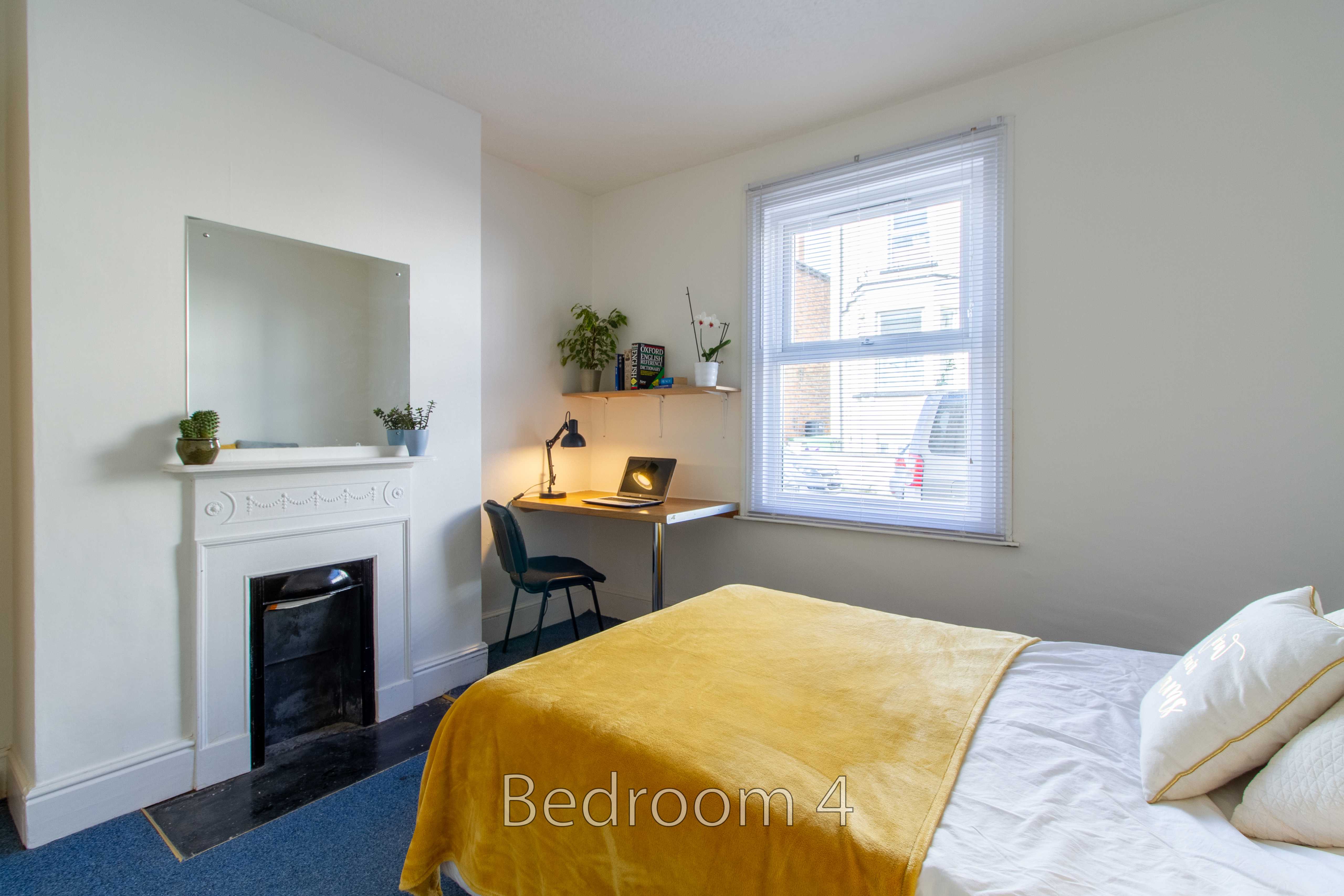 A bright bedroom with a double bed, yellow blanket, fireplace, desk, chair and window.