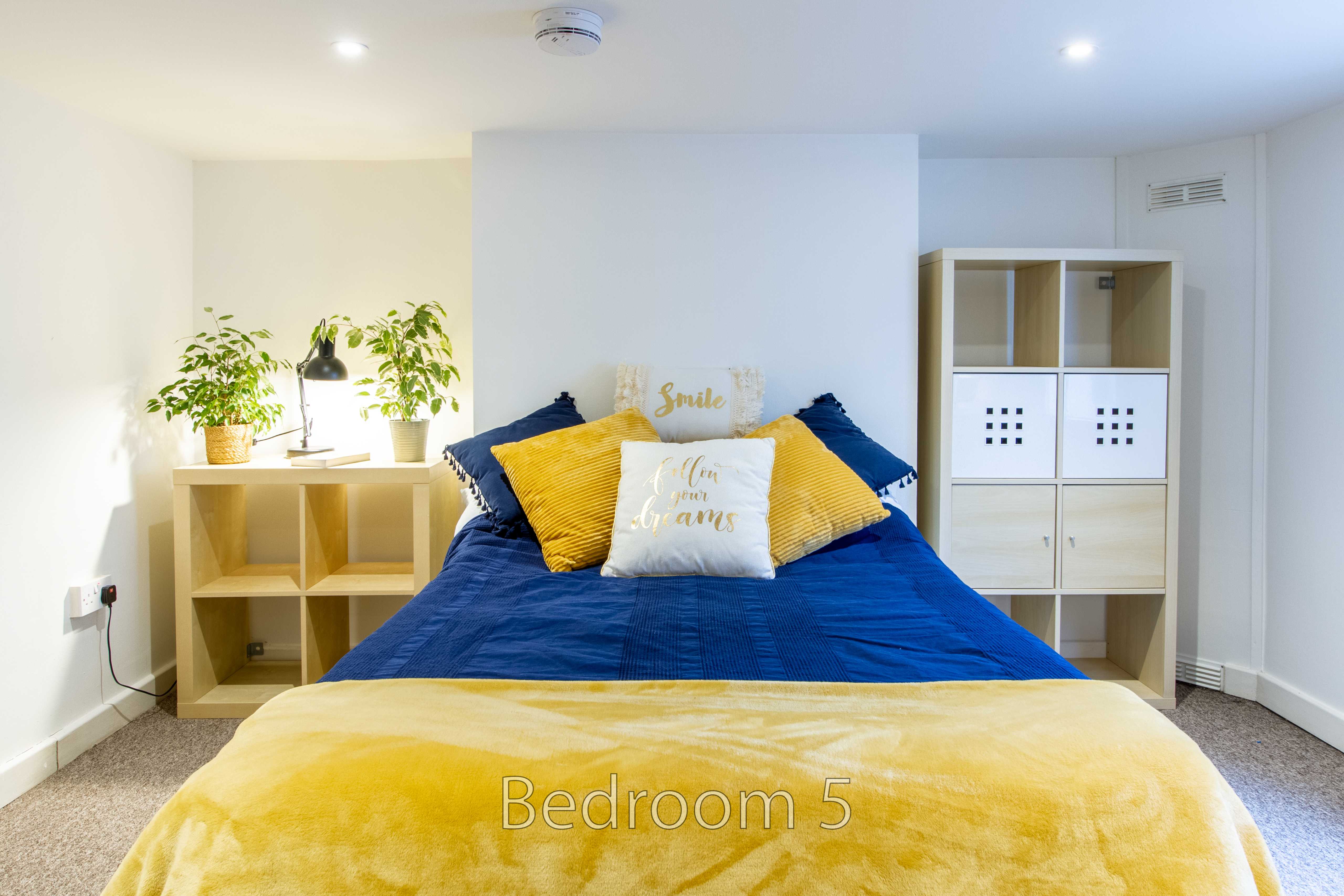 A furnished bedroom with a blue-covered bed, yellow blanket and pillows, shelving, plants and white walls.