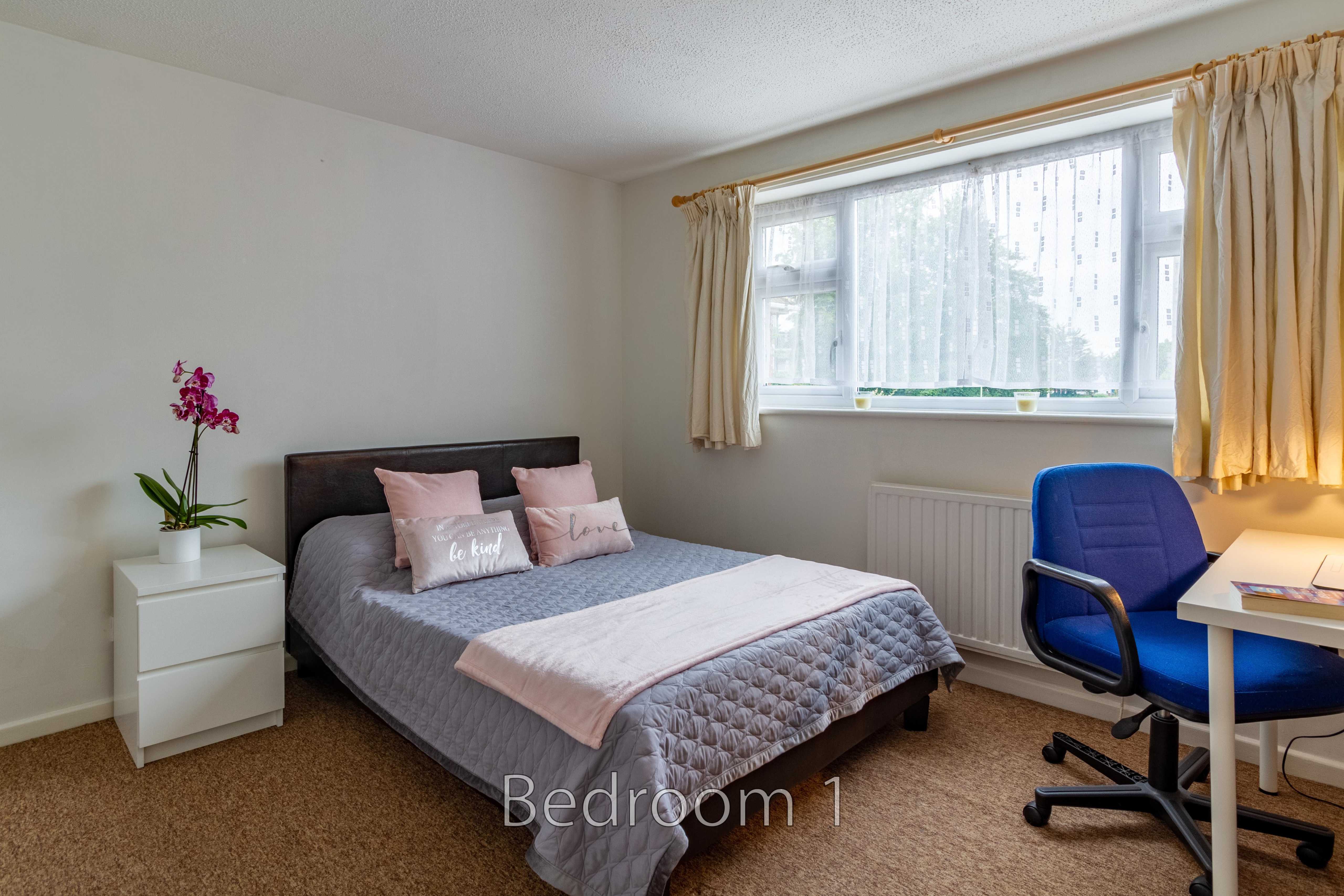 A furnished bedroom with a double bed, bedside table, window, curtains and a desk with an office chair.