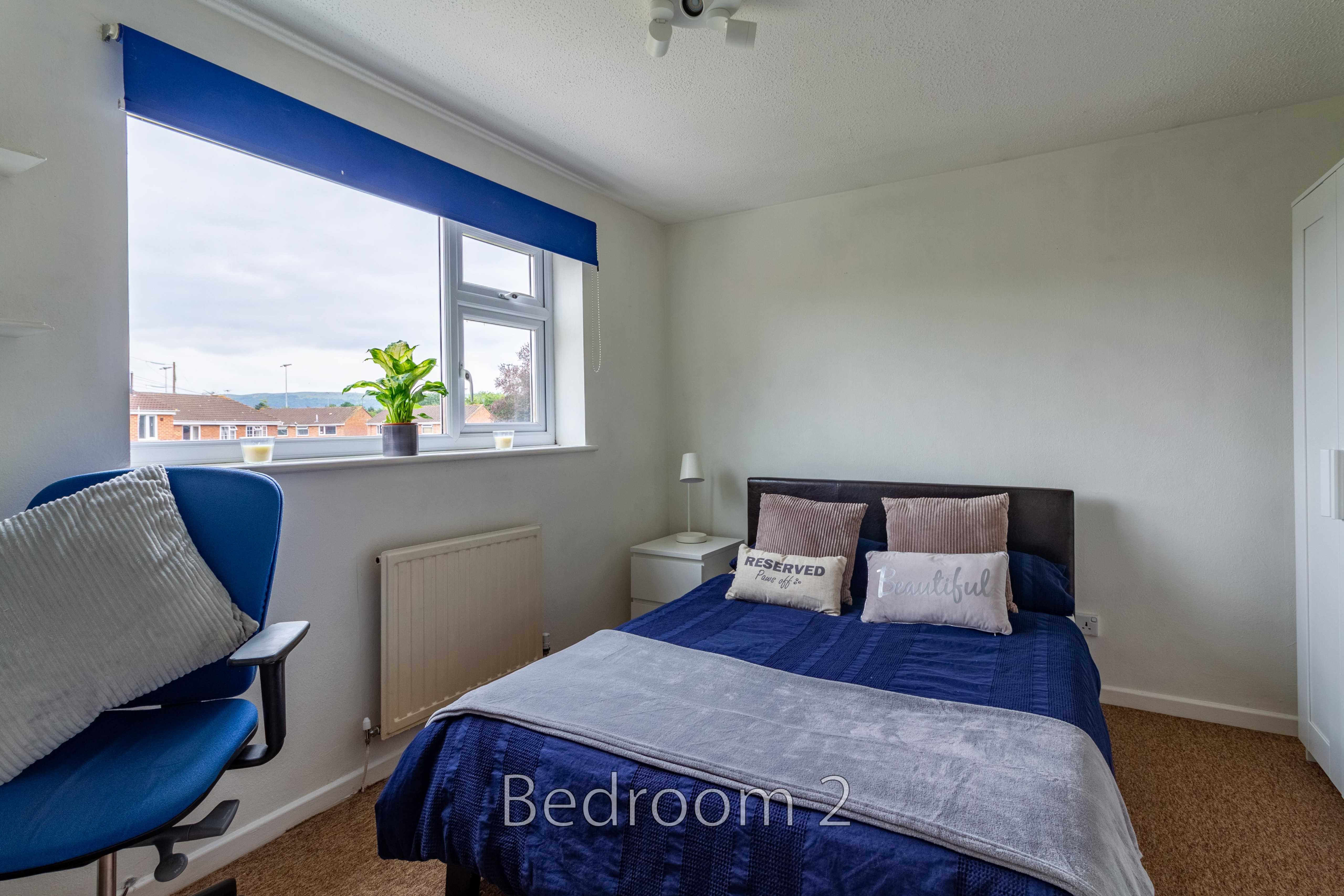 A bedroom with a double bed, blue bedding, a window, a desk chair and a wardrobe.