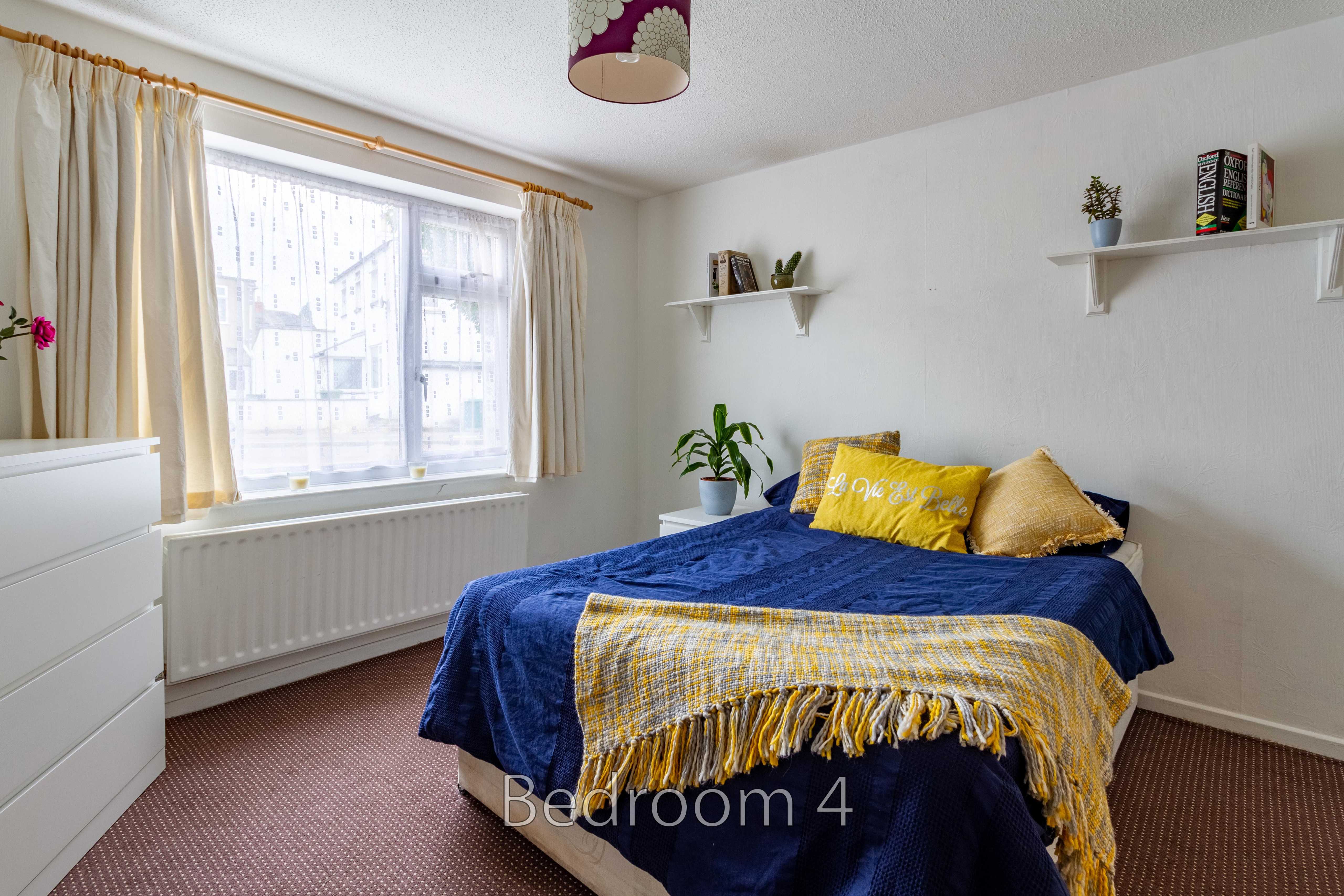 A furnished bedroom with a double bed, blue bedding, a window, radiator, chest of drawers and wall shelves.