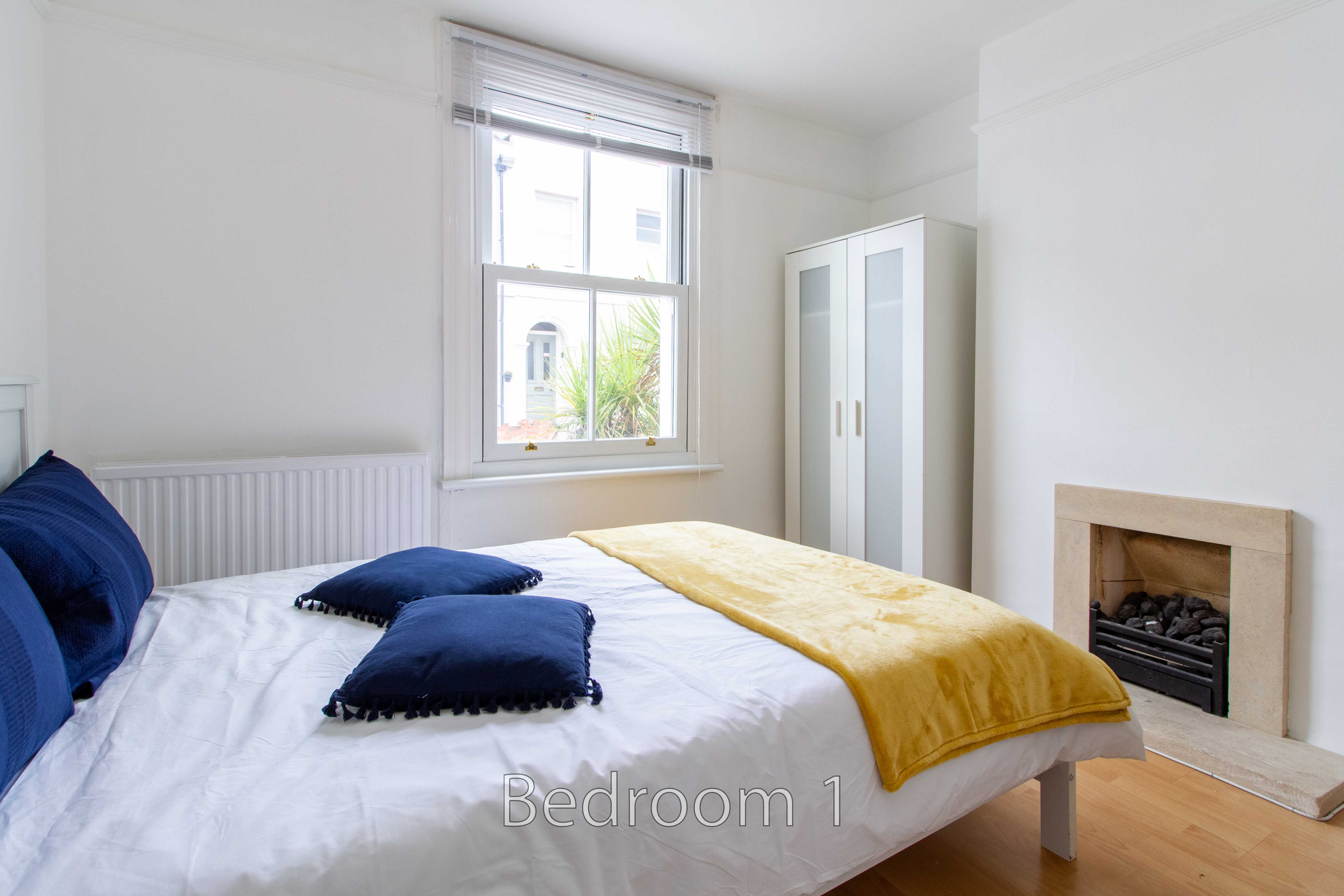 A bright bedroom with a bed, blue pillows, a yellow throw, wardrobe, window and fireplace.