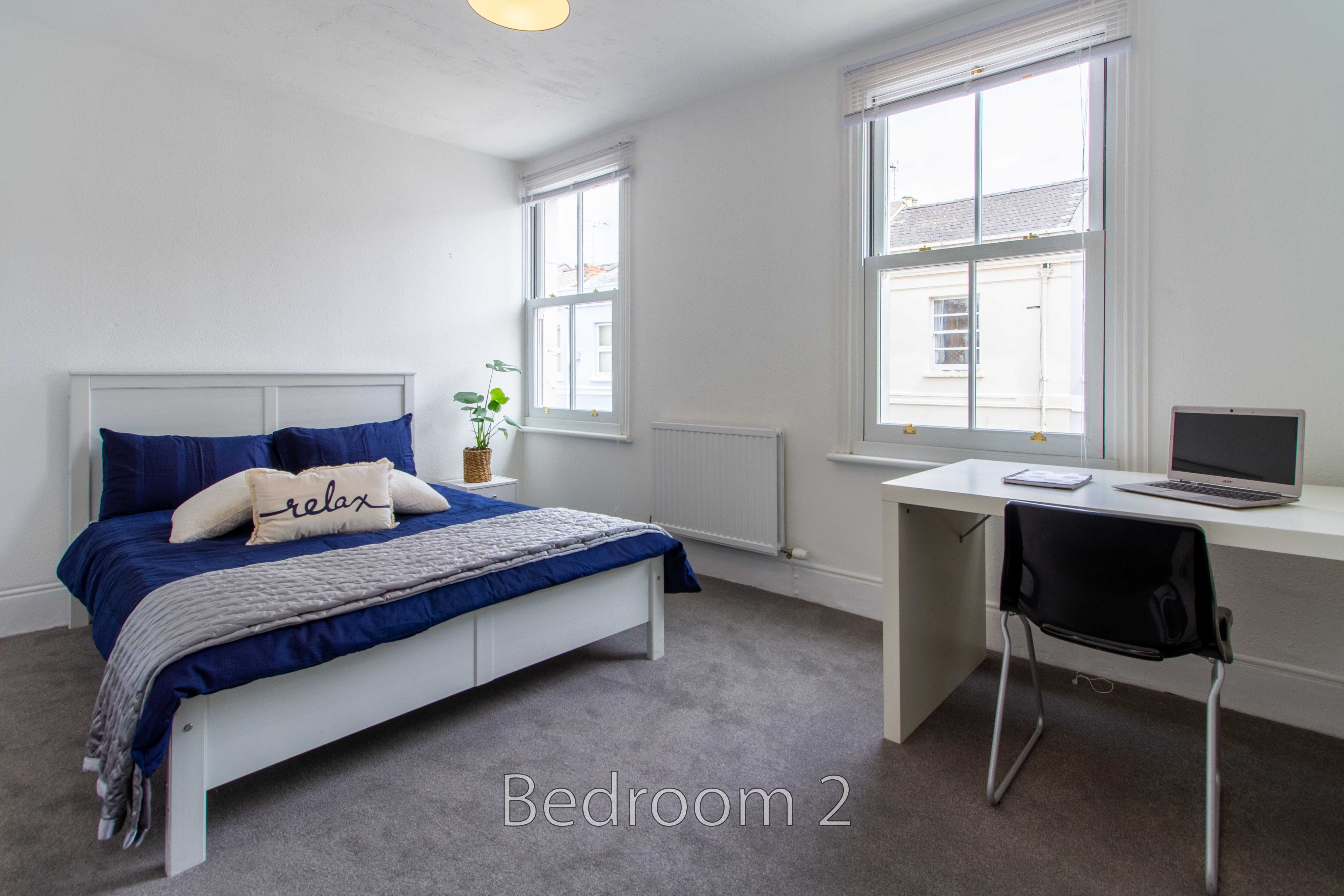 A furnished bedroom with a bed, desk, chair, two windows and a radiator.