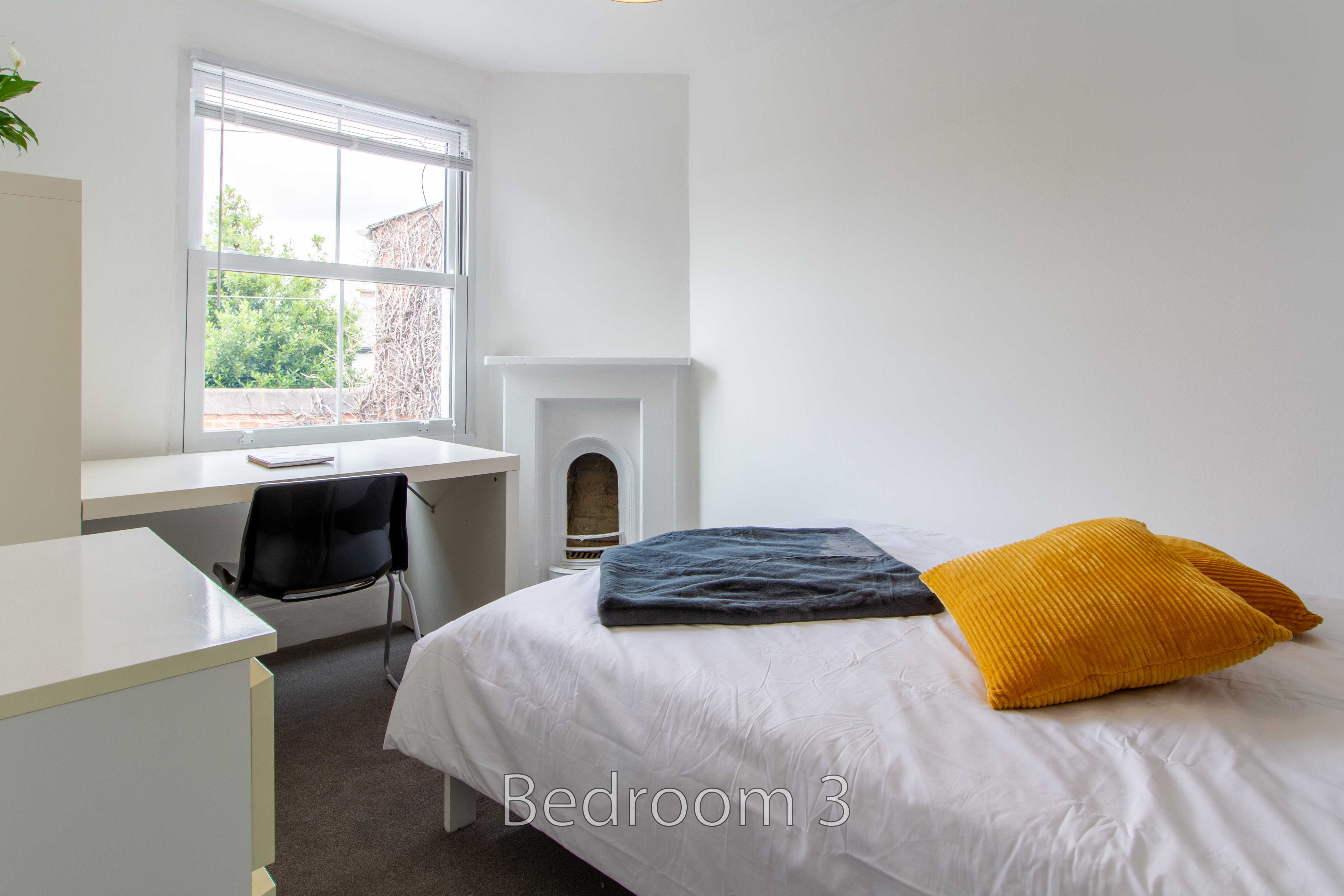 A bright bedroom with a double bed, desk, chair, window and fireplace.
