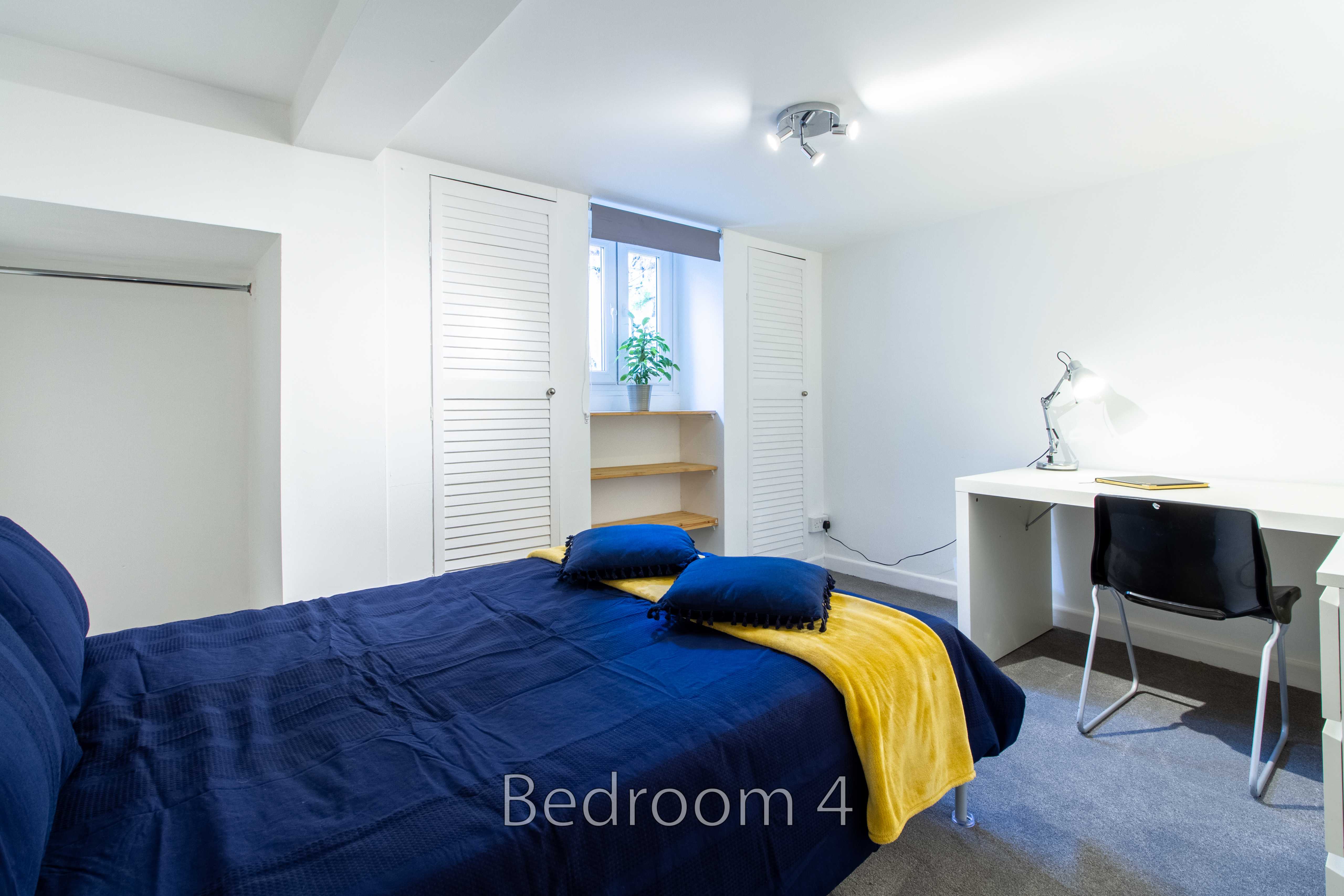 A bright bedroom with a blue bed, yellow throw, desk and chair, wardrobe doors, and a window.