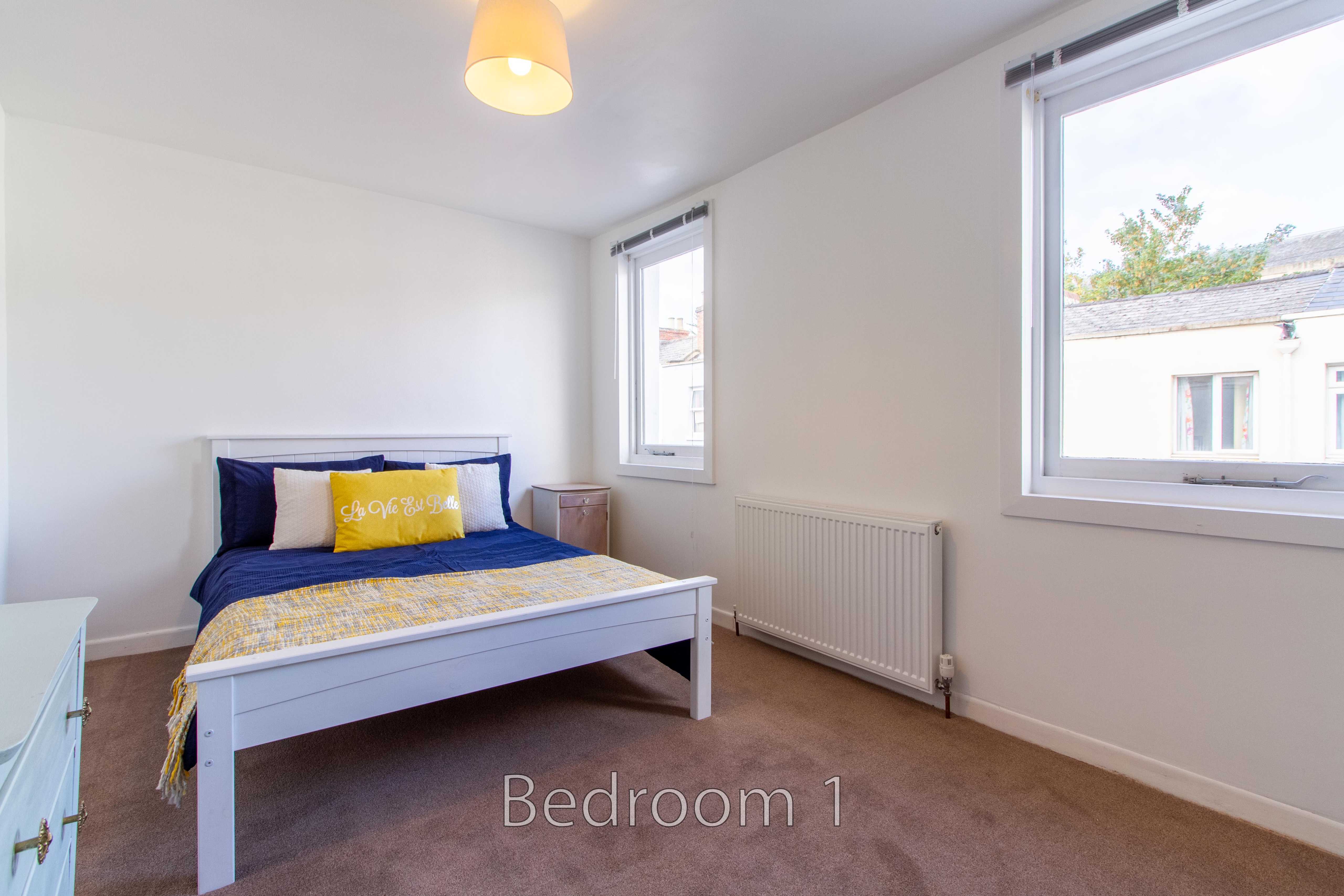 A bright bedroom with a double bed, two windows, a radiator and carpeted floor.