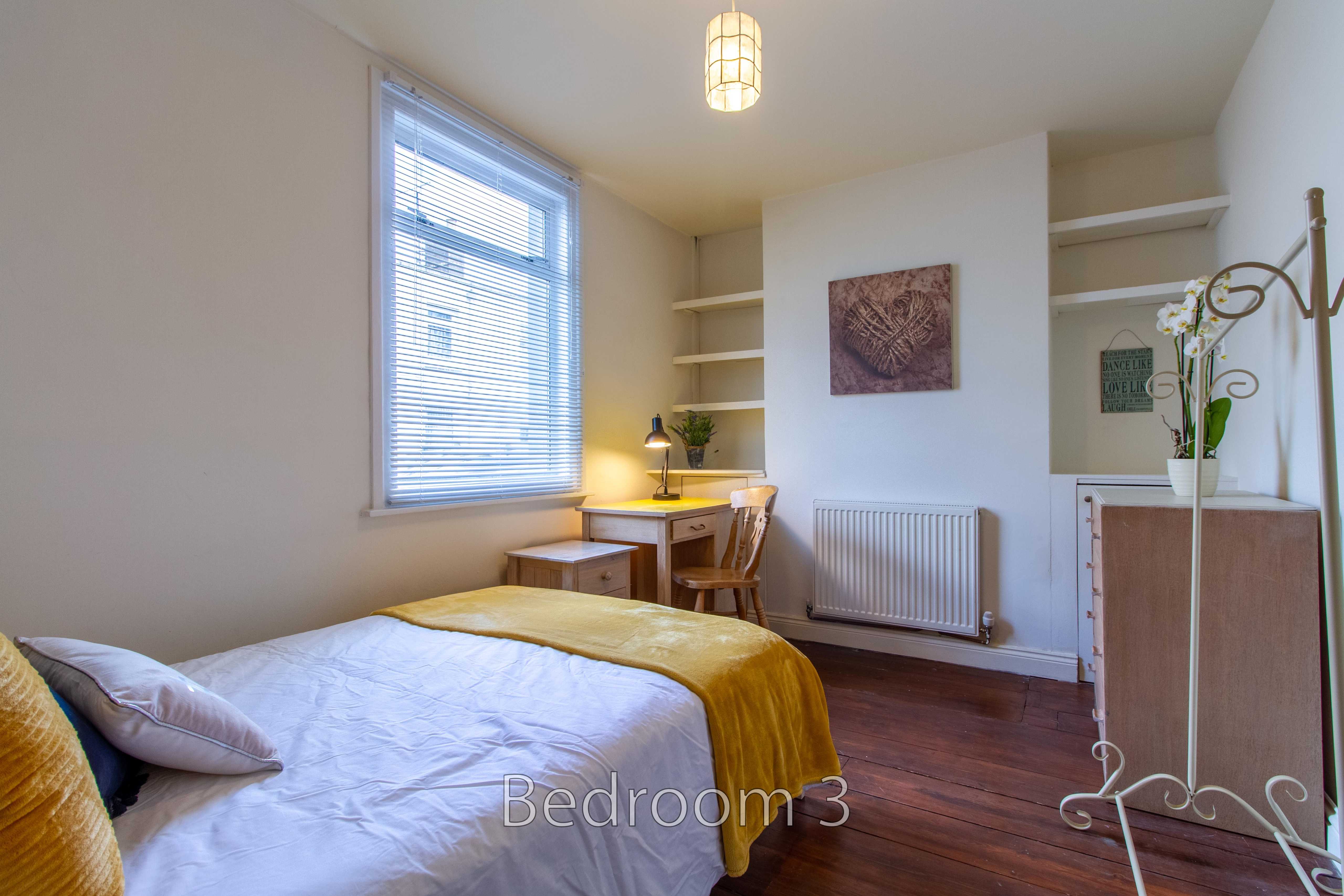 A furnished bedroom with a bed, desk, chair, window blinds, radiator and built-in shelving.