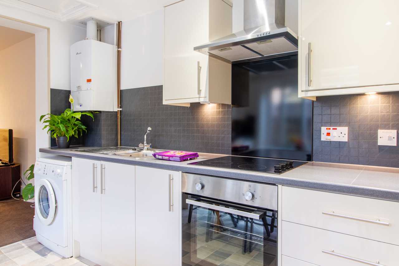 Modern fitted kitchen with white cabinets, oven, hob, extractor hood, washing machine and tiled backsplash.